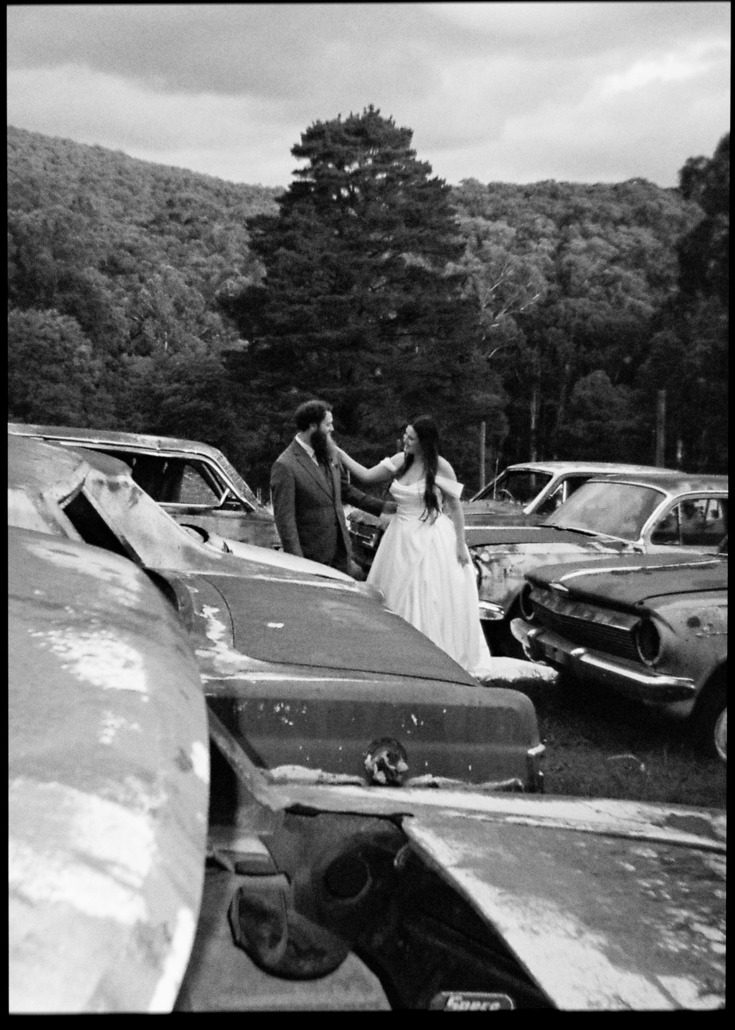 Black and white photo of a bride and groom posing romantically among rusty abandoned cars in a junkyard.