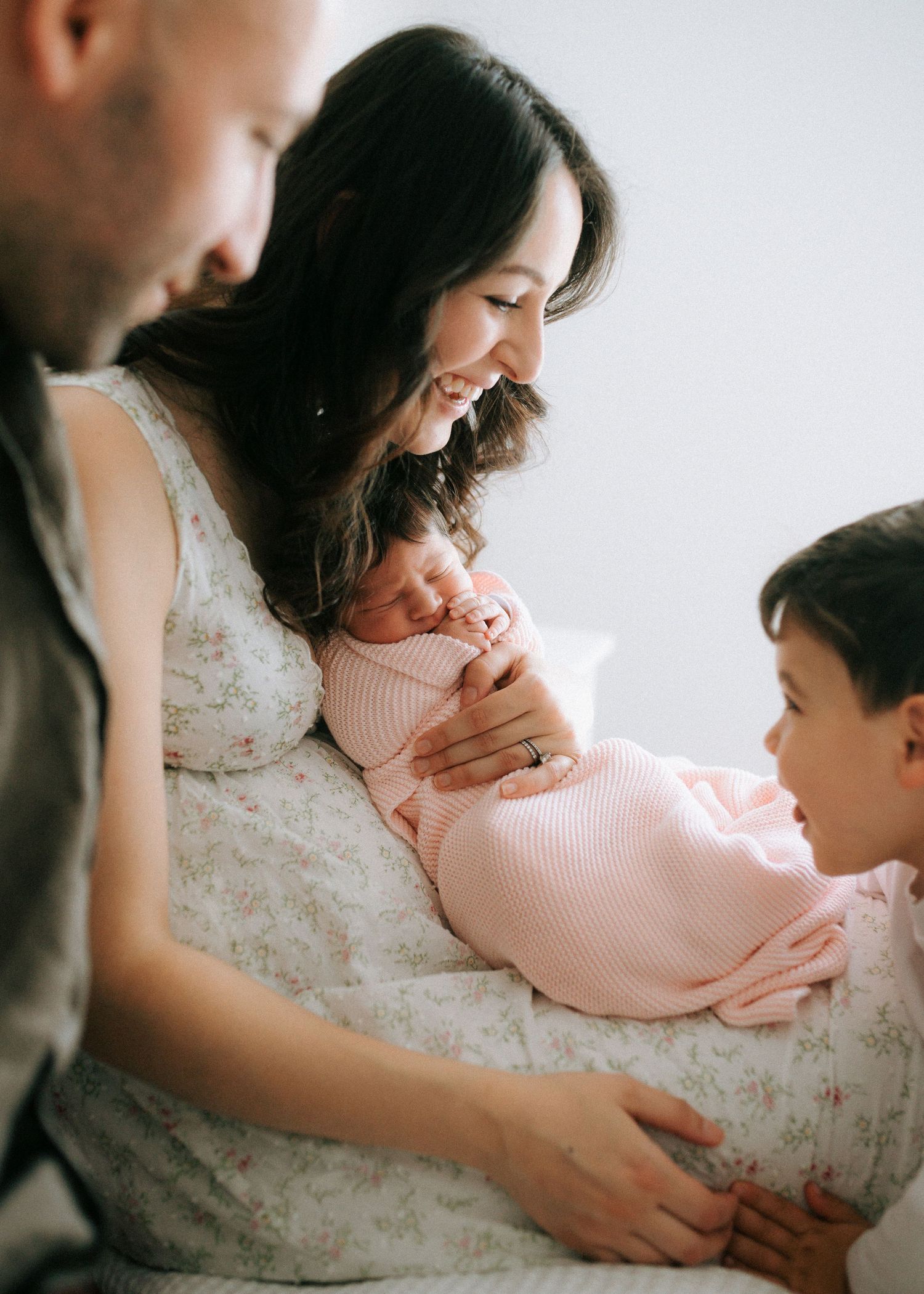 Family of four with father, smiling mother, toddler son, and sleeping newborn in pink swaddle.