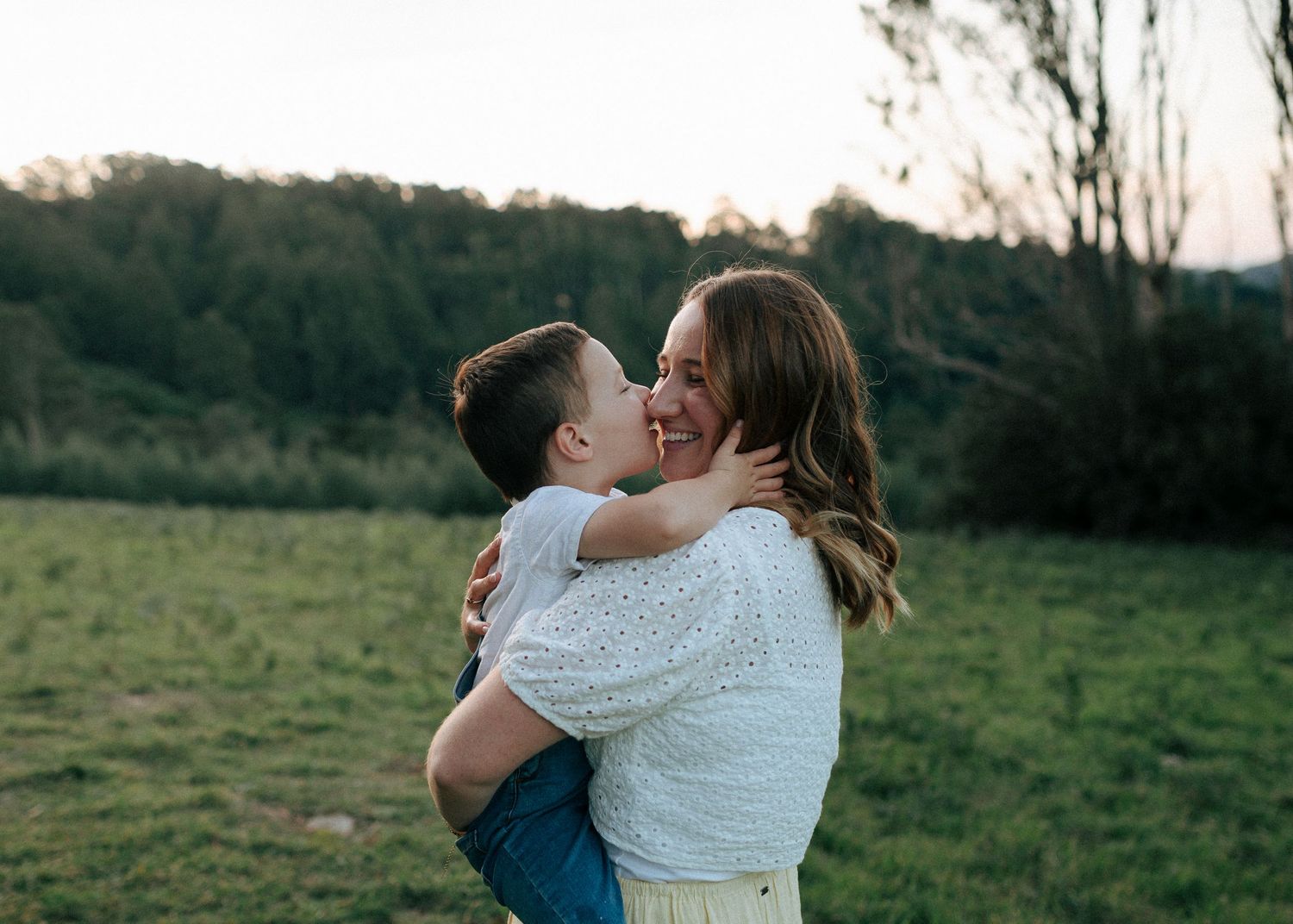 A mother laughs joyfully as her young son gives her a sweet kiss on the cheek outdoors.