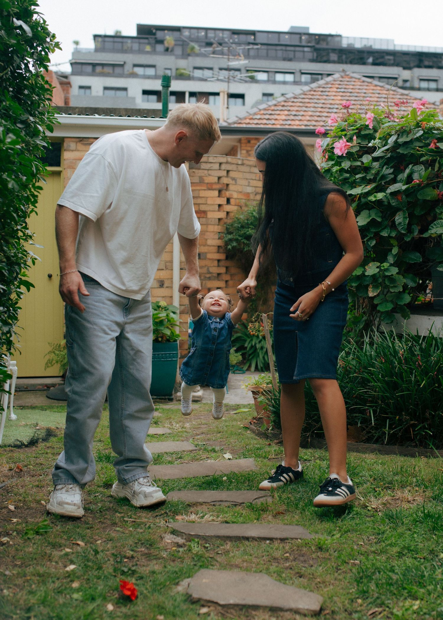 Parents holding toddler's hands while walking together in a green backyard garden.