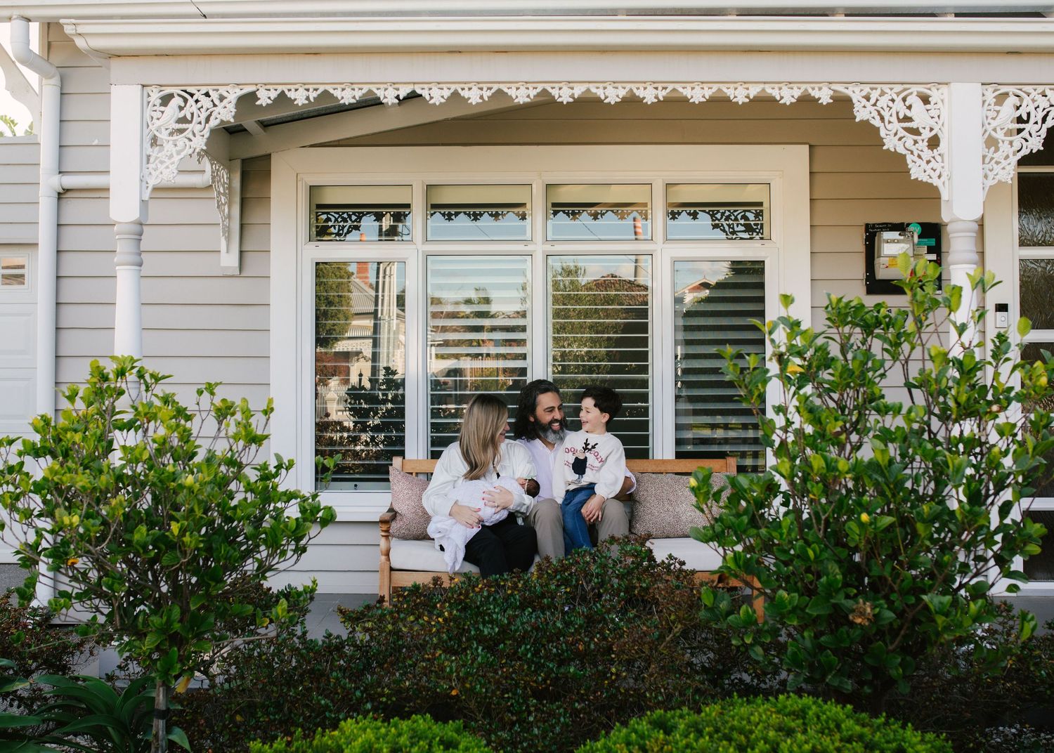 Young family sitting together on front porch of beautiful white heritage cottage with gardens.