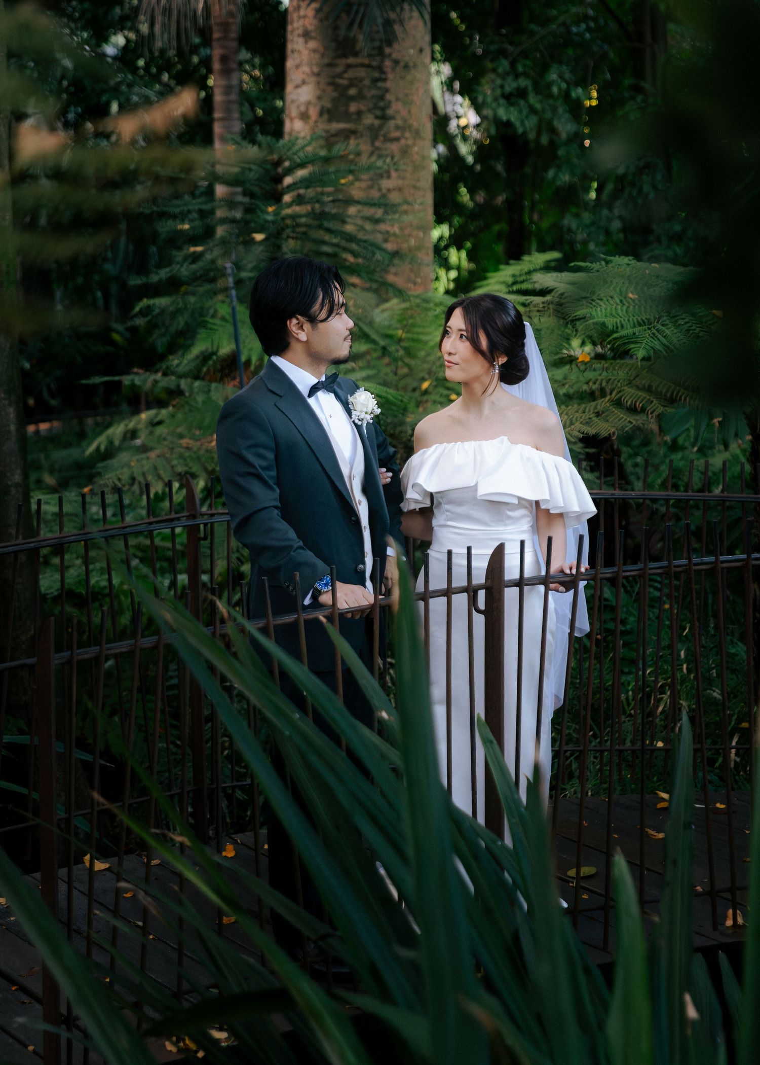 Bride and groom stand on a garden bridge surrounded by lush green ferns and trees, gazing at each other.