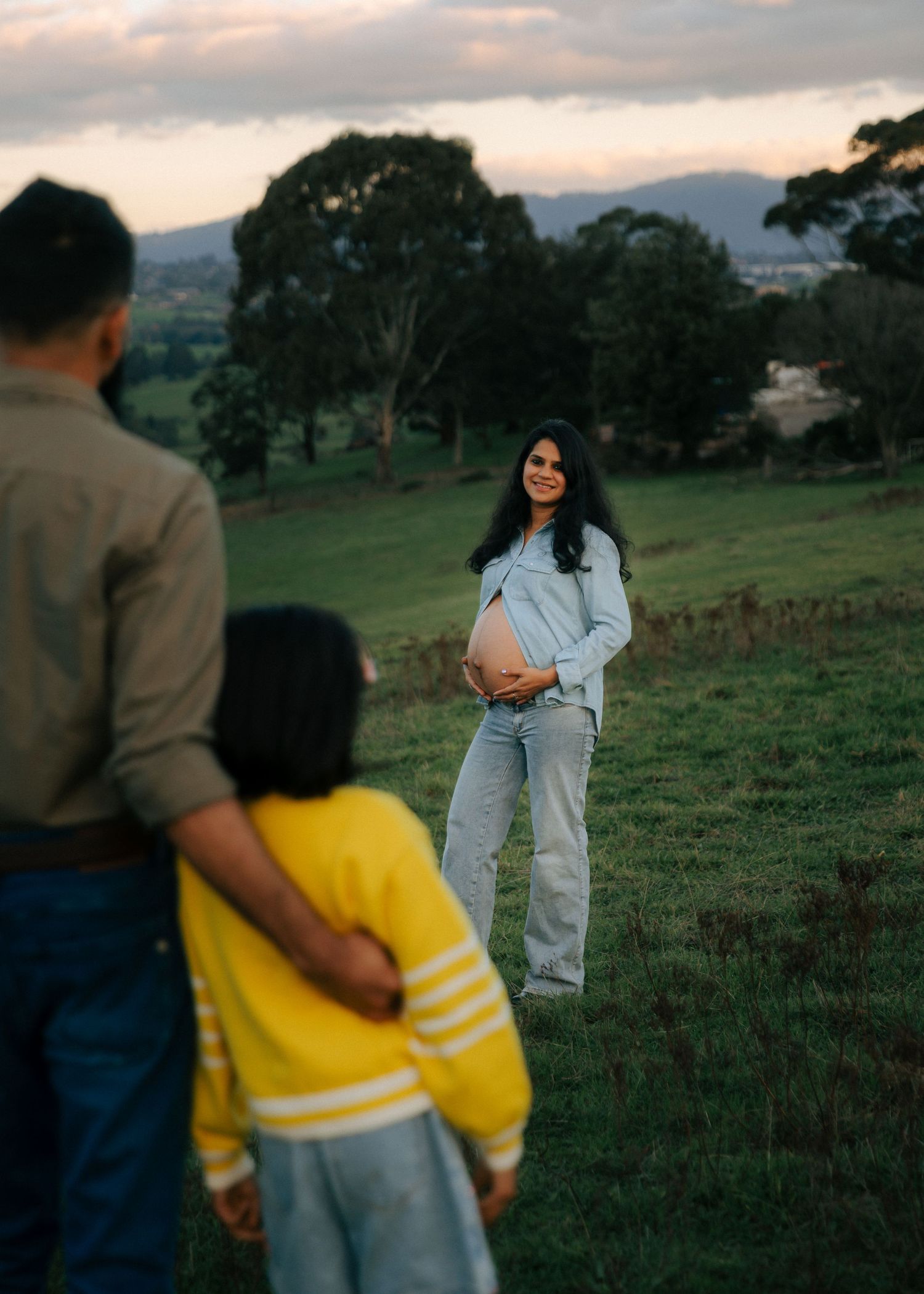 Pregnant woman smiling and holding baby bump as partner and child in yellow sweater approach on green field.