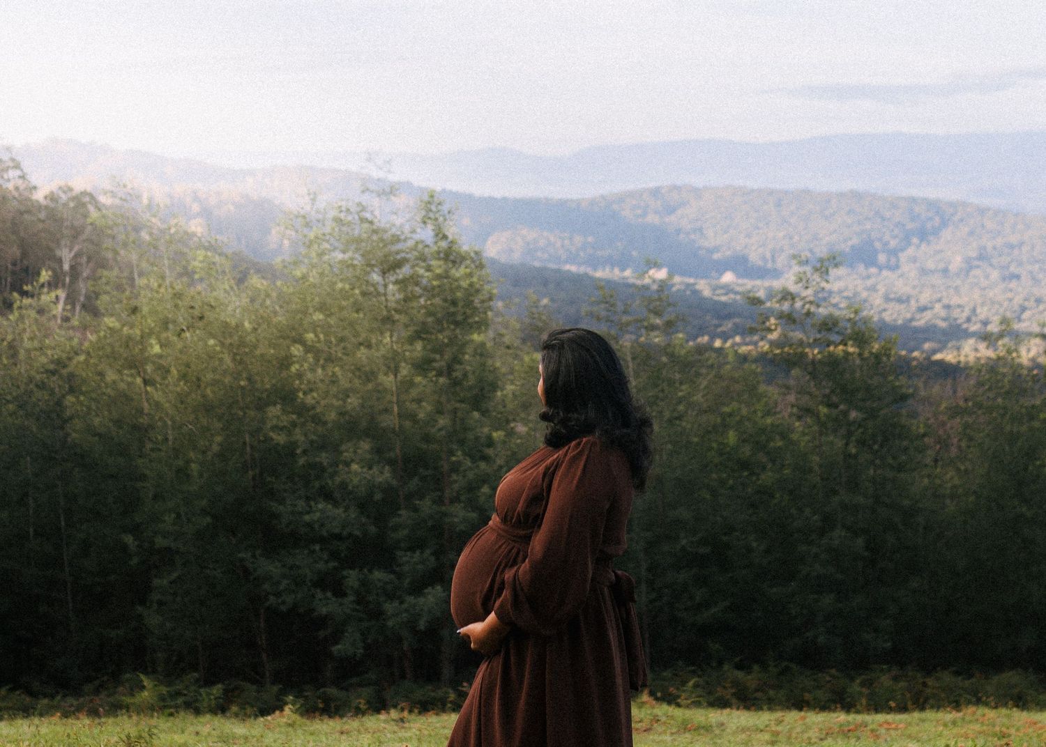 Pregnant woman in a flowing burgundy dress cradles her belly while standing in a grassy meadow with forested mountains in the background.