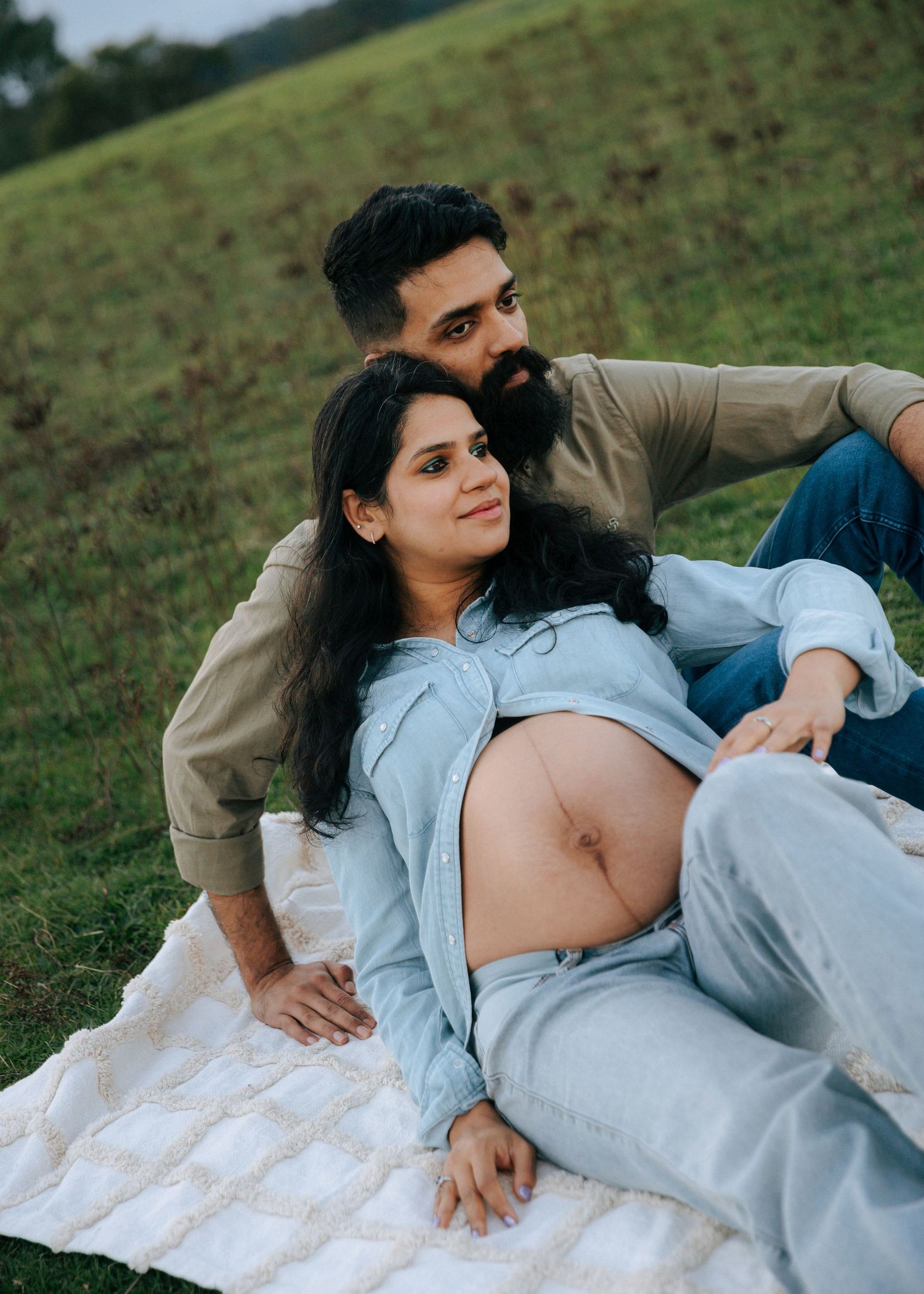 Couple sharing tender maternity photo moment on blanket in green field, woman showing pregnant belly.