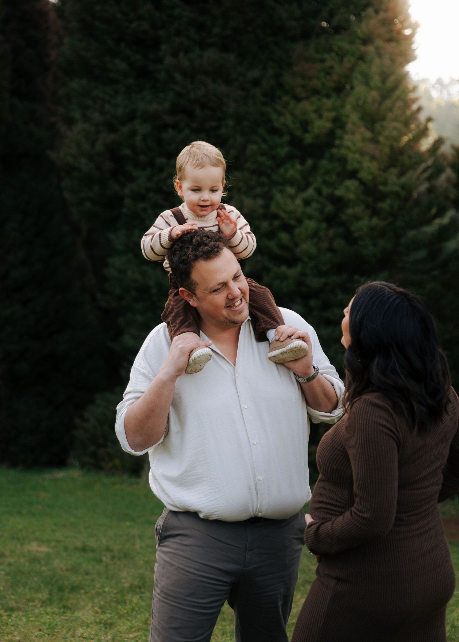 Father lifts laughing toddler onto shoulders while pregnant wife looks on in green park.