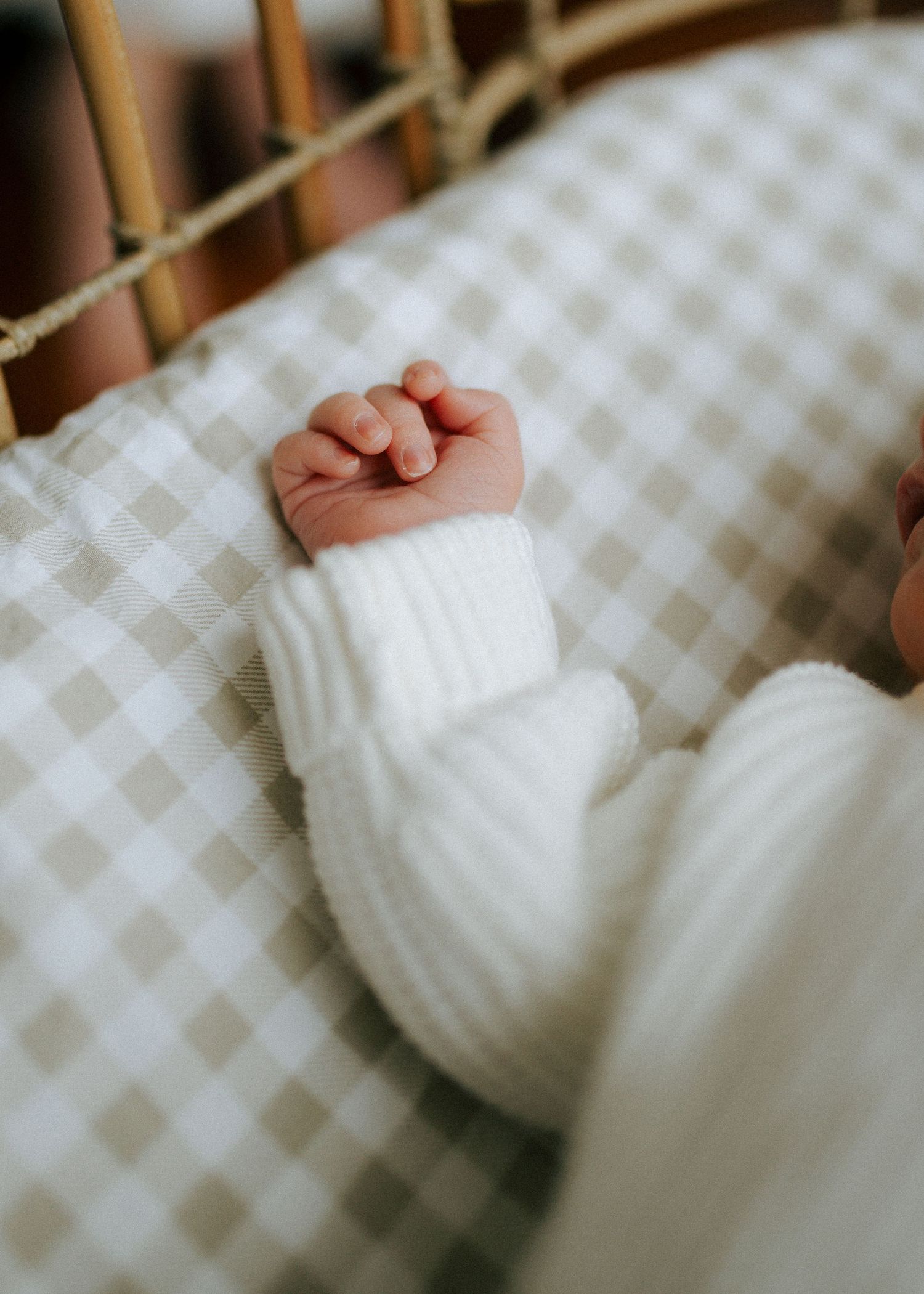 Close-up of newborn baby tiny clenched fist in white ribbed knit sleeve on gingham bassinet sheet.