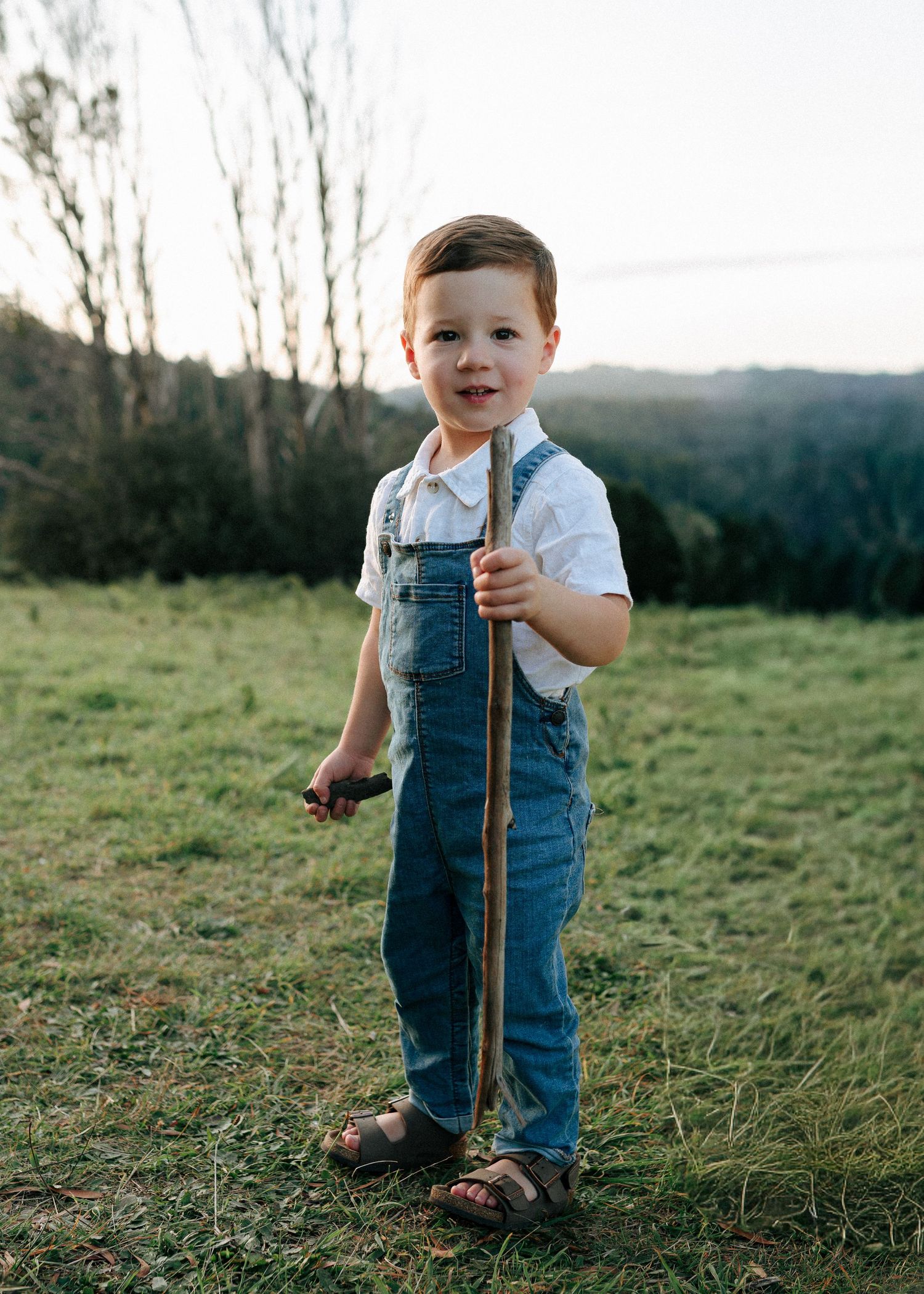 An adorable toddler boy in denim overalls holds a stick while standing in a grassy field.