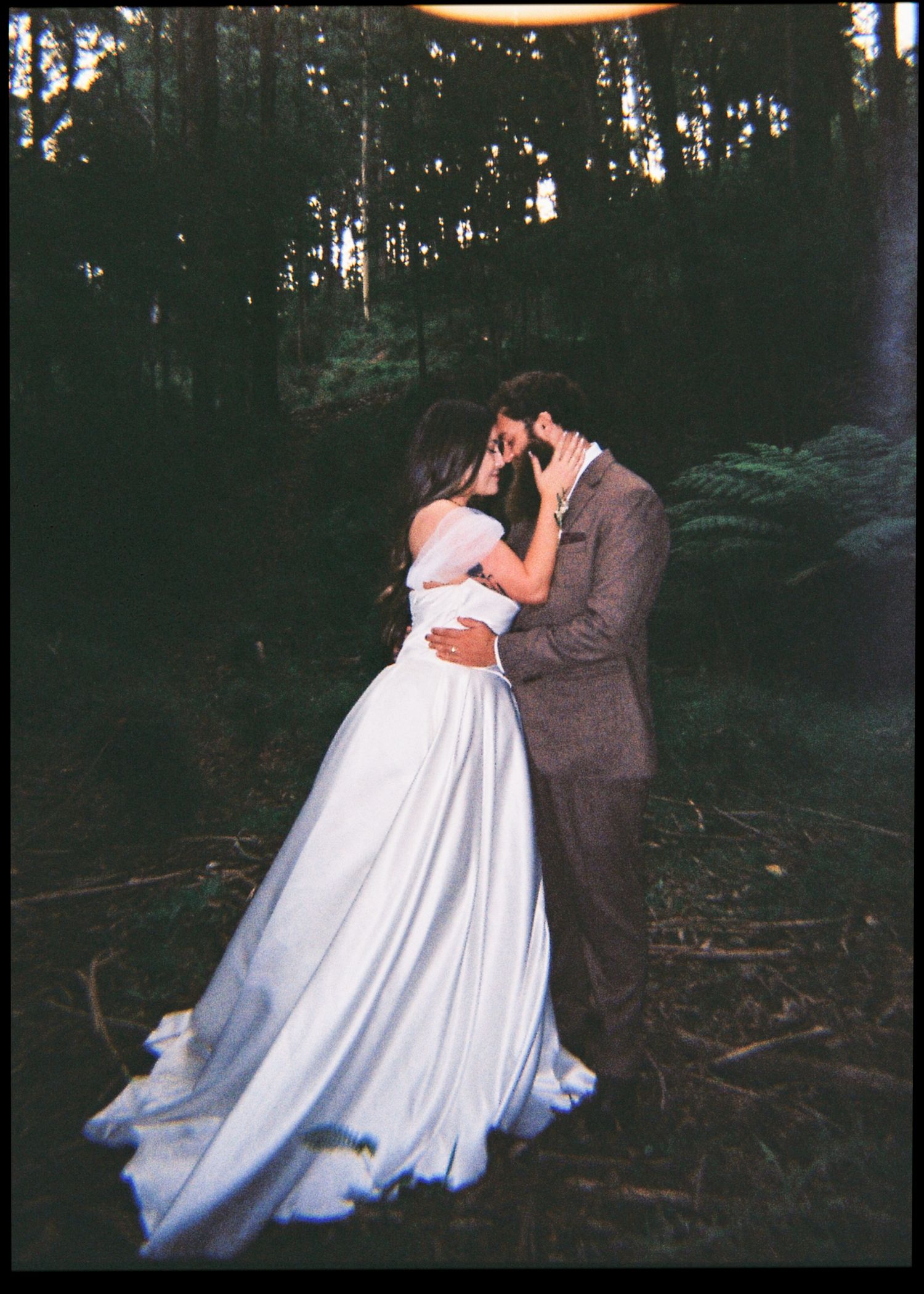 Bride and groom sharing a romantic kiss in a moody forest setting, bride in elegant white ball gown.