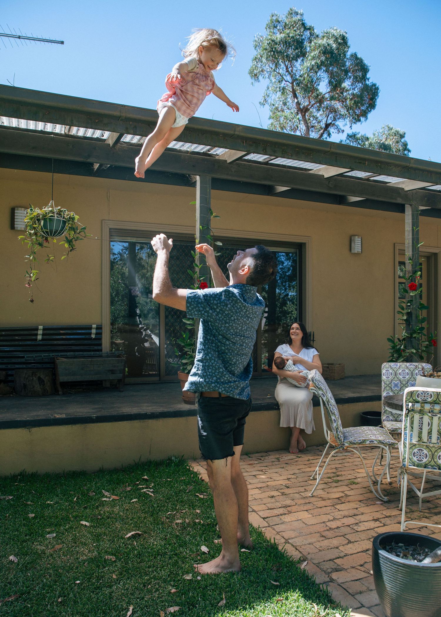 A father tosses his young child in the air in a backyard while a mother sits holding a baby nearby.