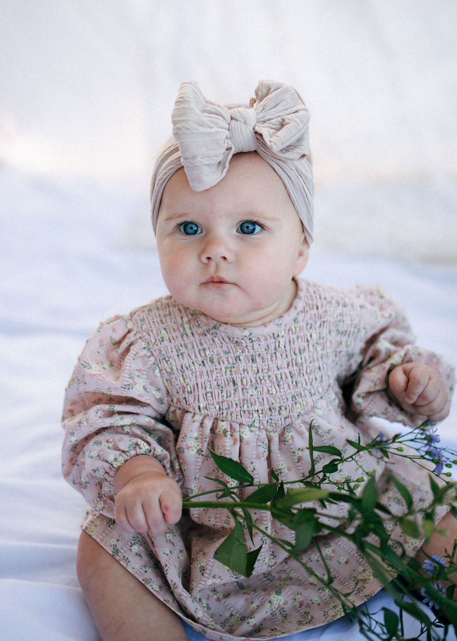 Blue-eyed baby girl in floral dress and bow headband with greenery.