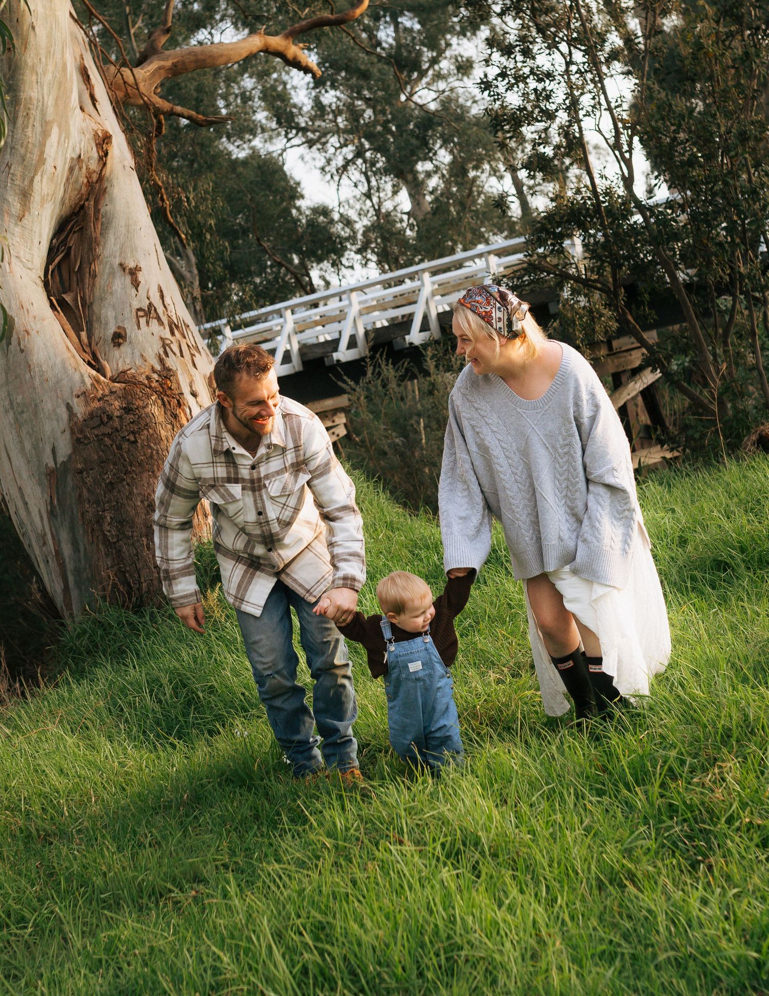Family of three walking on lush green grass near large eucalyptus tree outdoors.