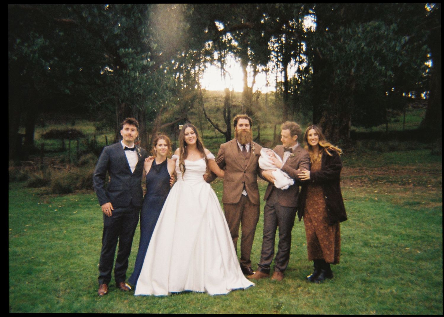 Wedding party group portrait at sunset in a lush green garden, bride in white gown surrounded by family and friends.