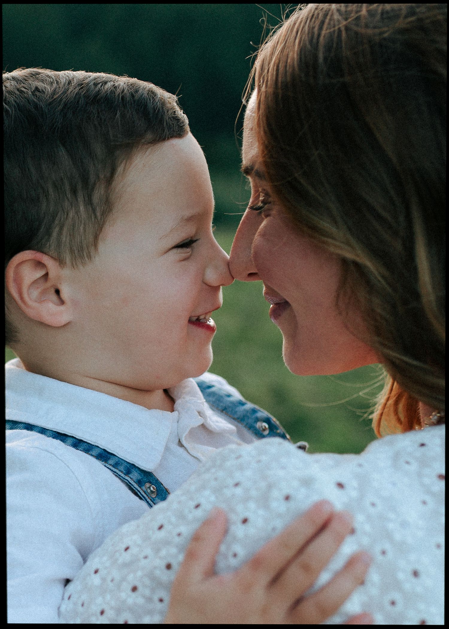 A smiling young boy and his mother touch noses in a tender close-up outdoor portrait.