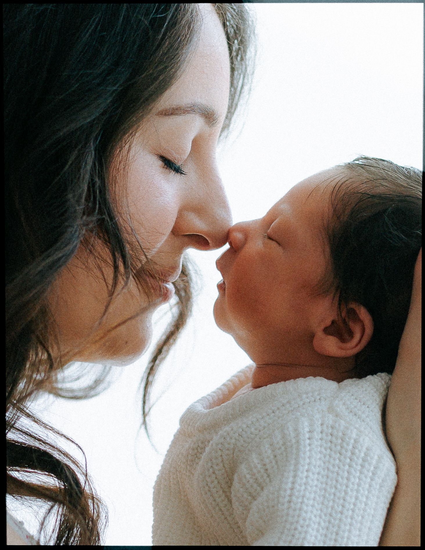 A dark-haired mother tenderly touches noses with her newborn baby in soft light.