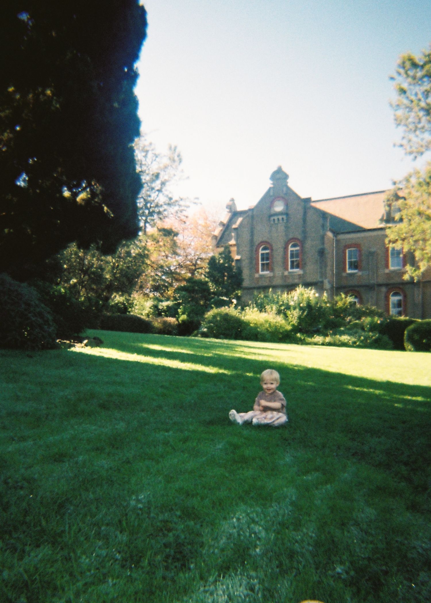 A baby sits alone on a green lawn in front of a large historic stone manor building.