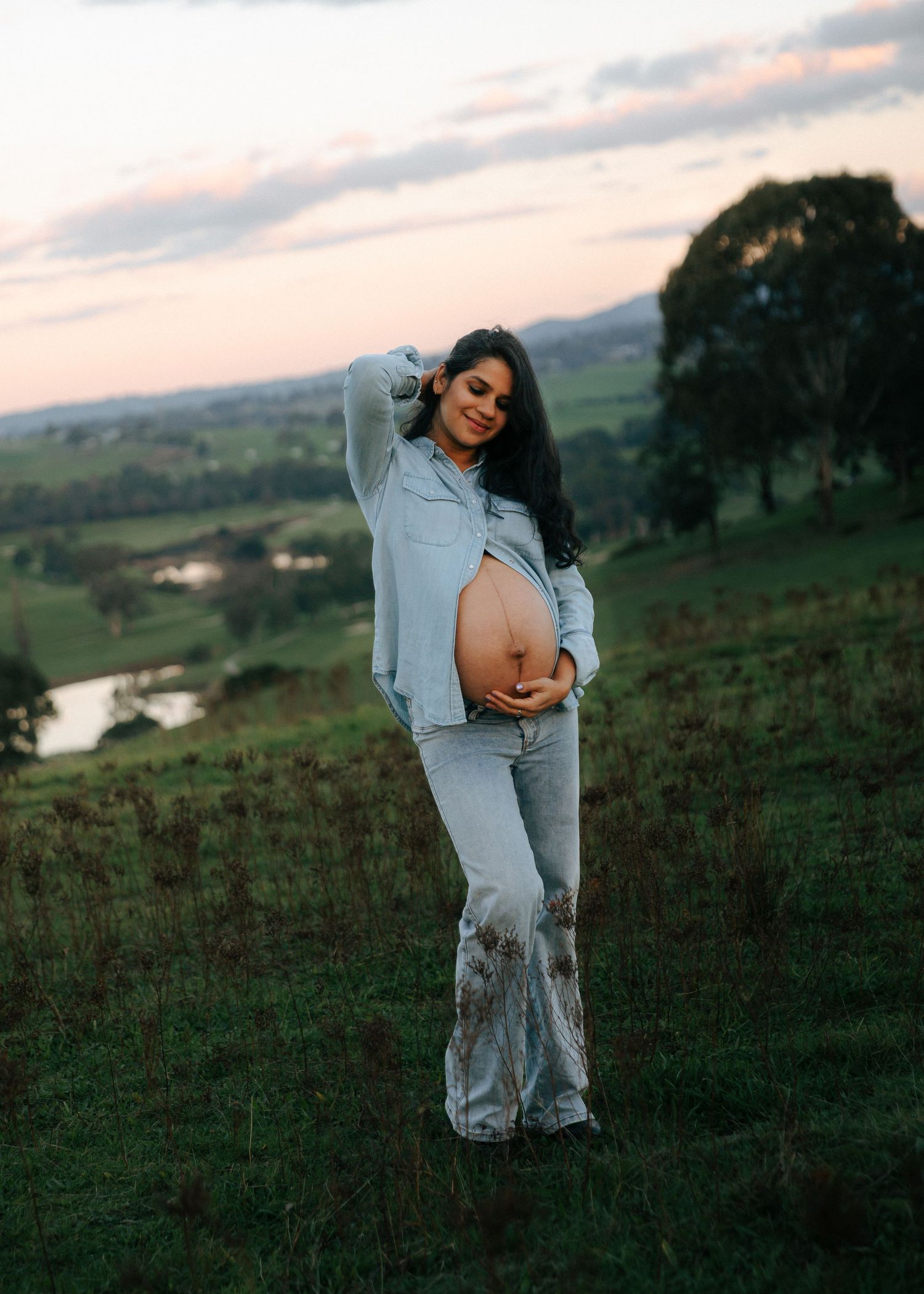 Glowing pregnant woman in denim outfit cradling her bare belly on a green hillside at sunset.