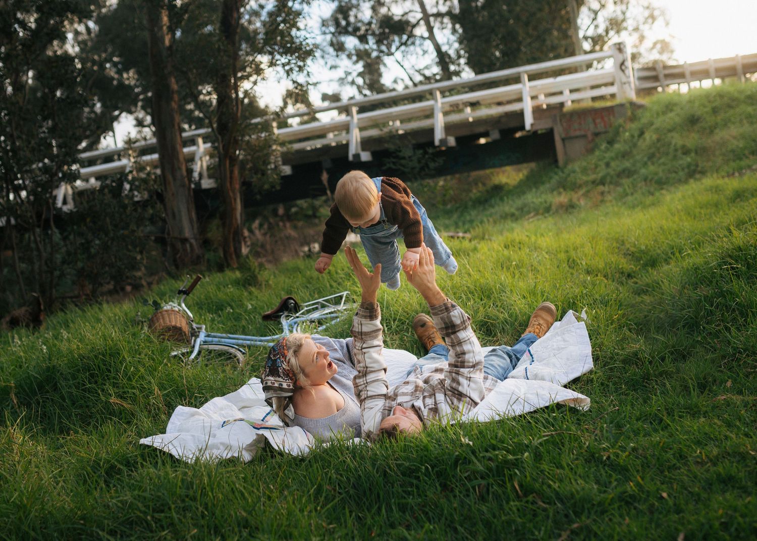 Mother lifting smiling toddler overhead while lying on a blanket in green grass near a bridge.