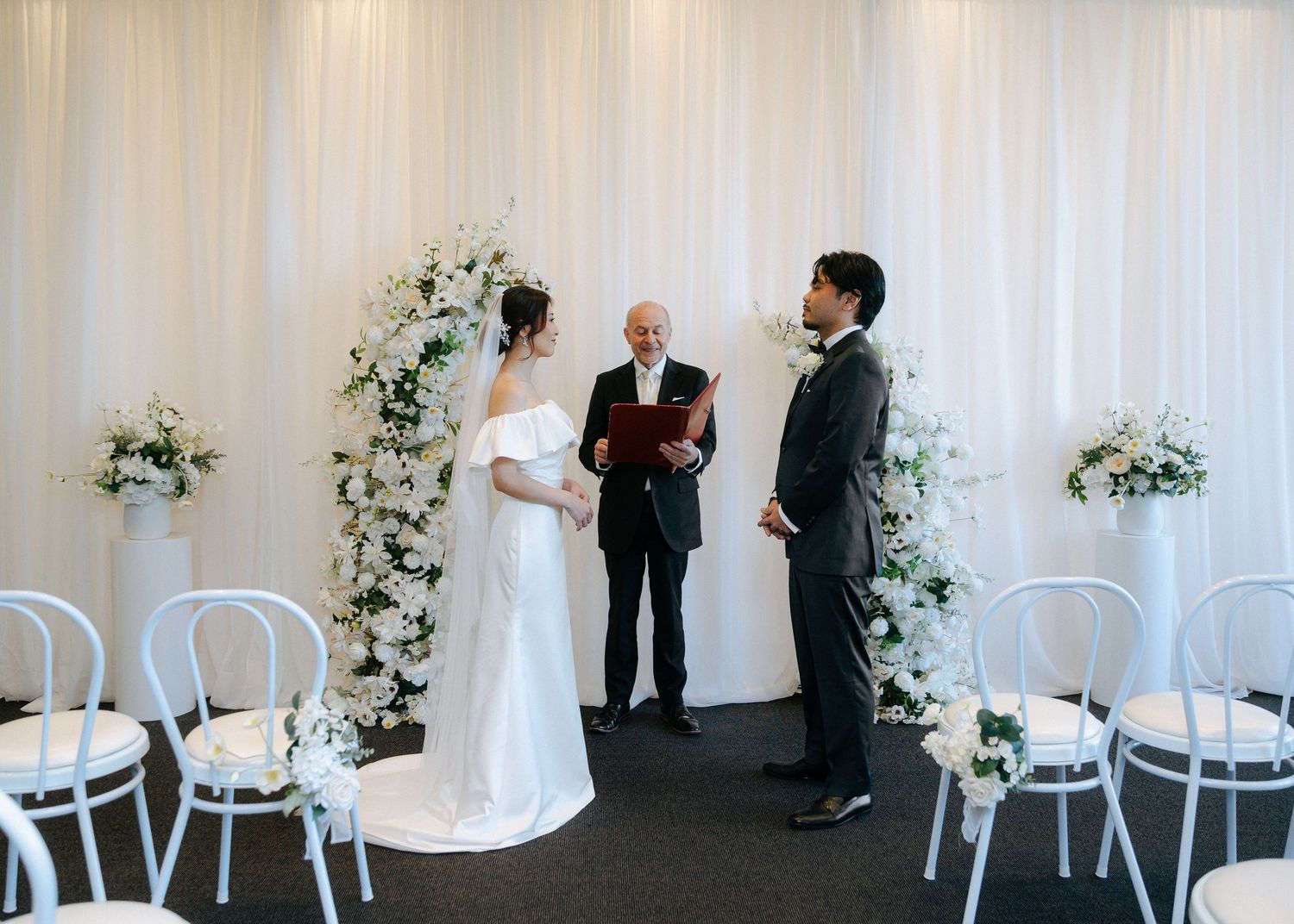Bride and groom exchanging vows before officiant at white draped altar with white floral pillars.