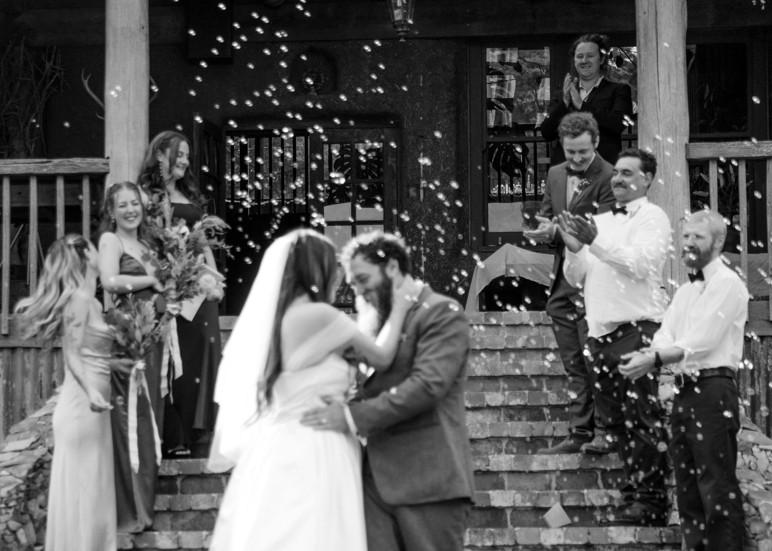 Black and white photo of newlyweds kissing on steps surrounded by guests throwing confetti.
