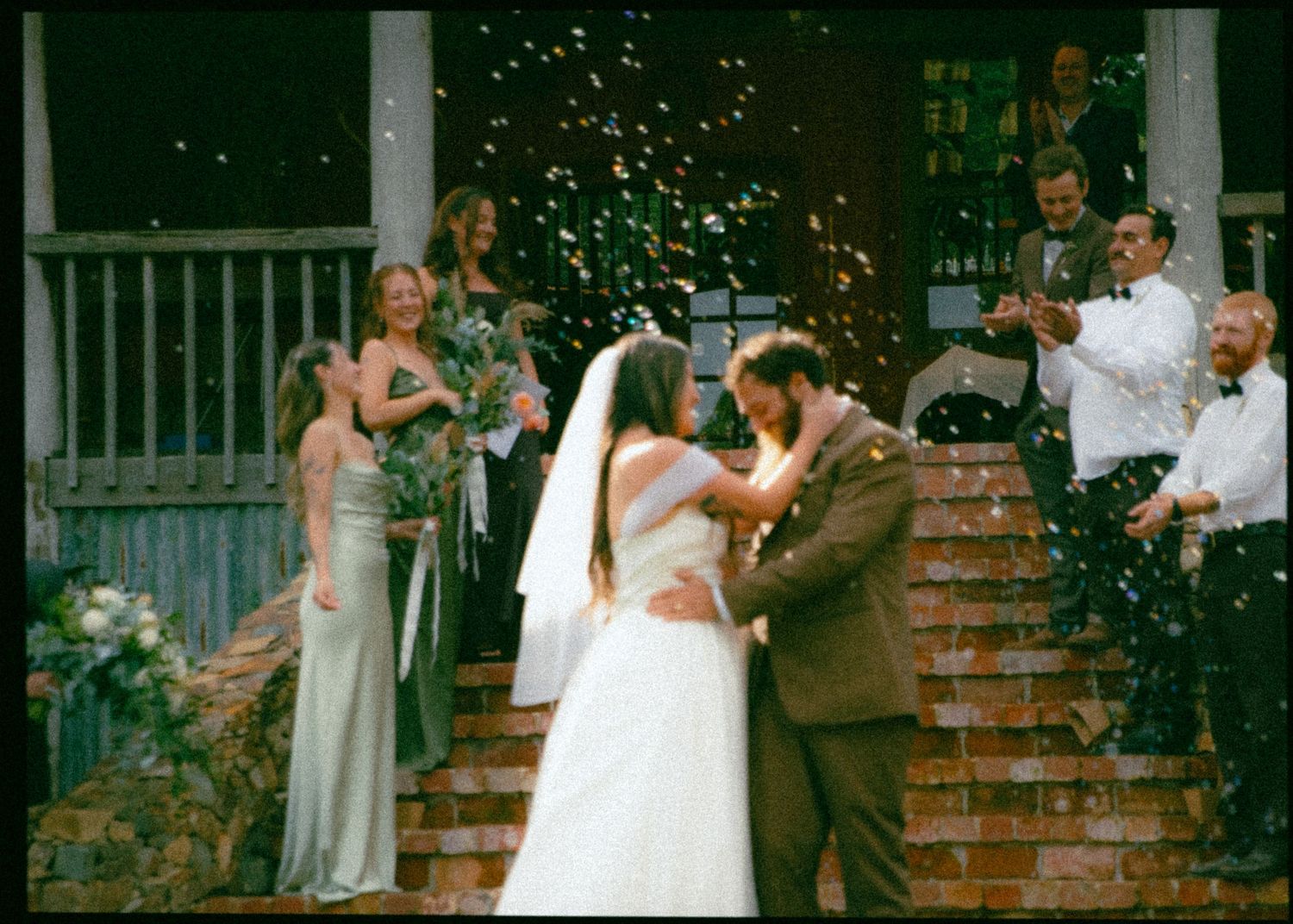 Bride and groom share a kiss on brick steps as guests toss confetti in celebration.