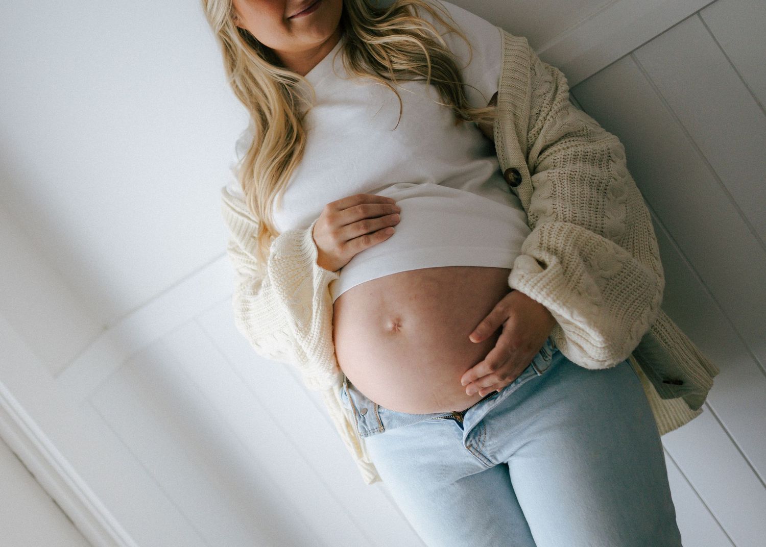 Close-up of pregnant woman in white crop top and jeans gently holding her bare baby bump.