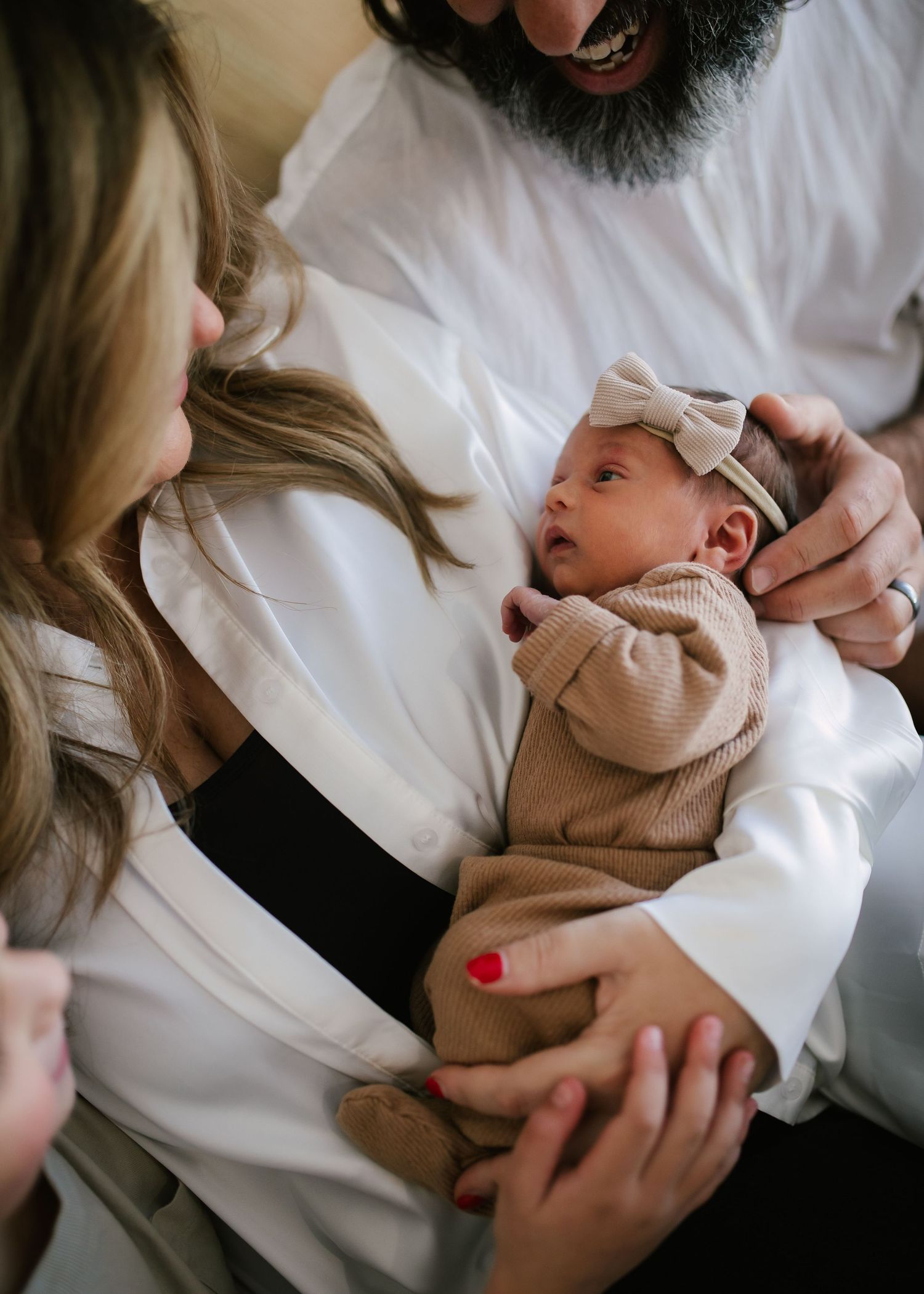 Parents admiring their newborn baby girl wearing a beige bow headband in their arms.