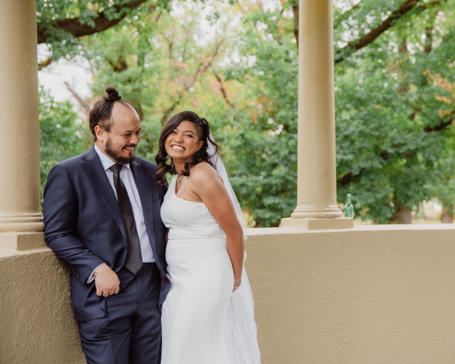 Bride and groom laughing together beside columns in a park pavilion setting.