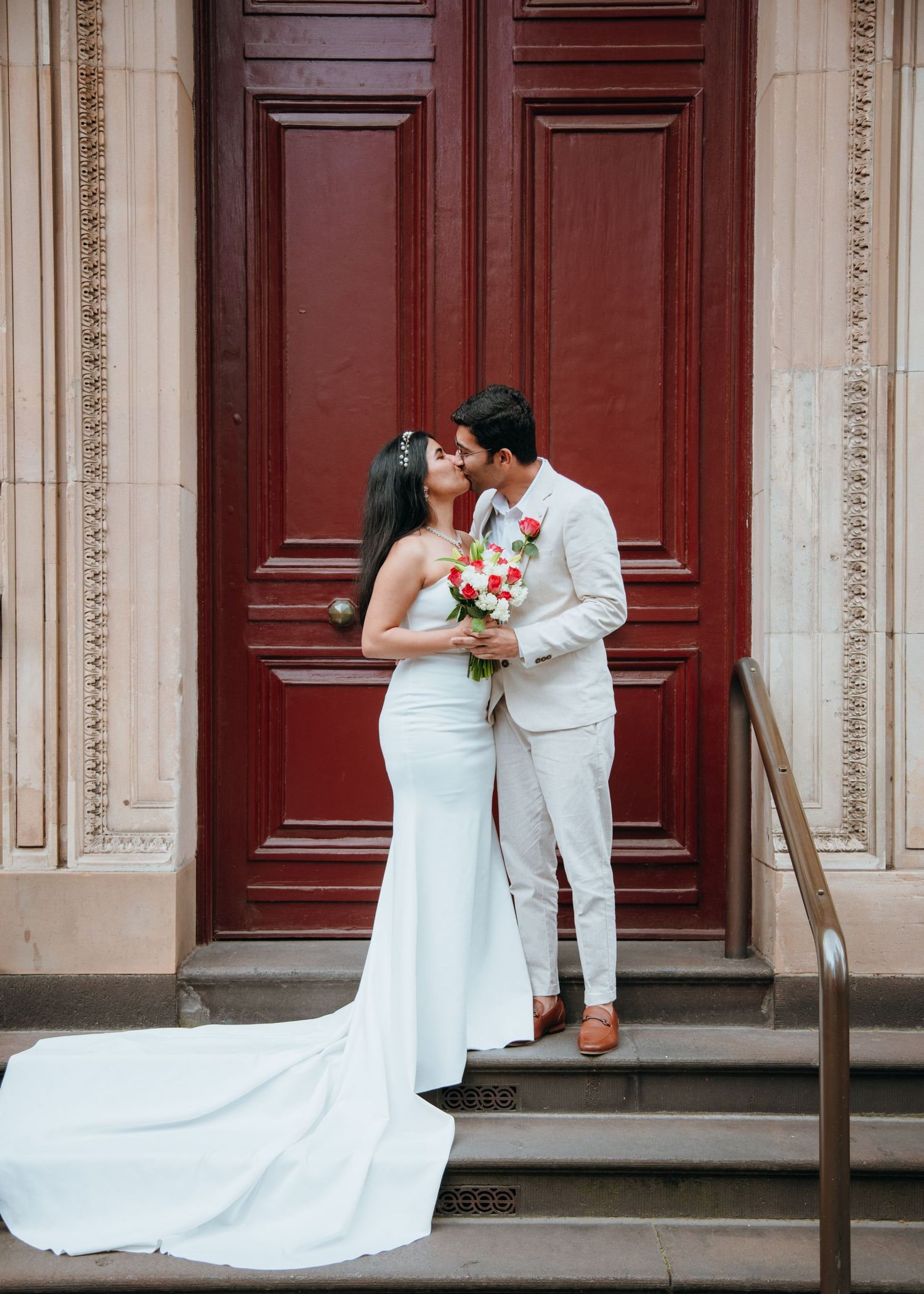 Bride and groom share first kiss holding red rose bouquet in front of large ornate red wooden door.