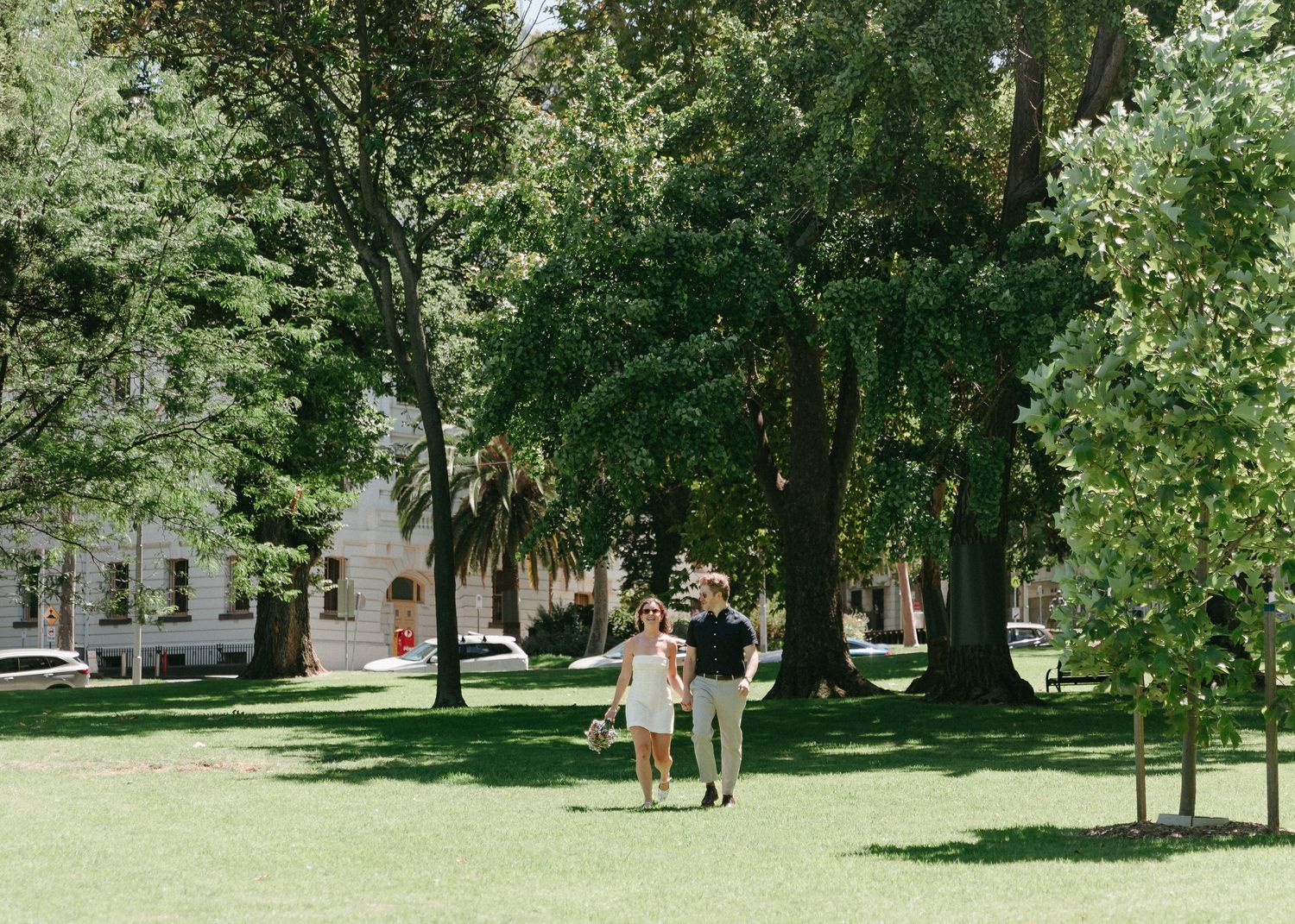 Newlywed couple walks hand in hand through lush green park on sunny wedding day.