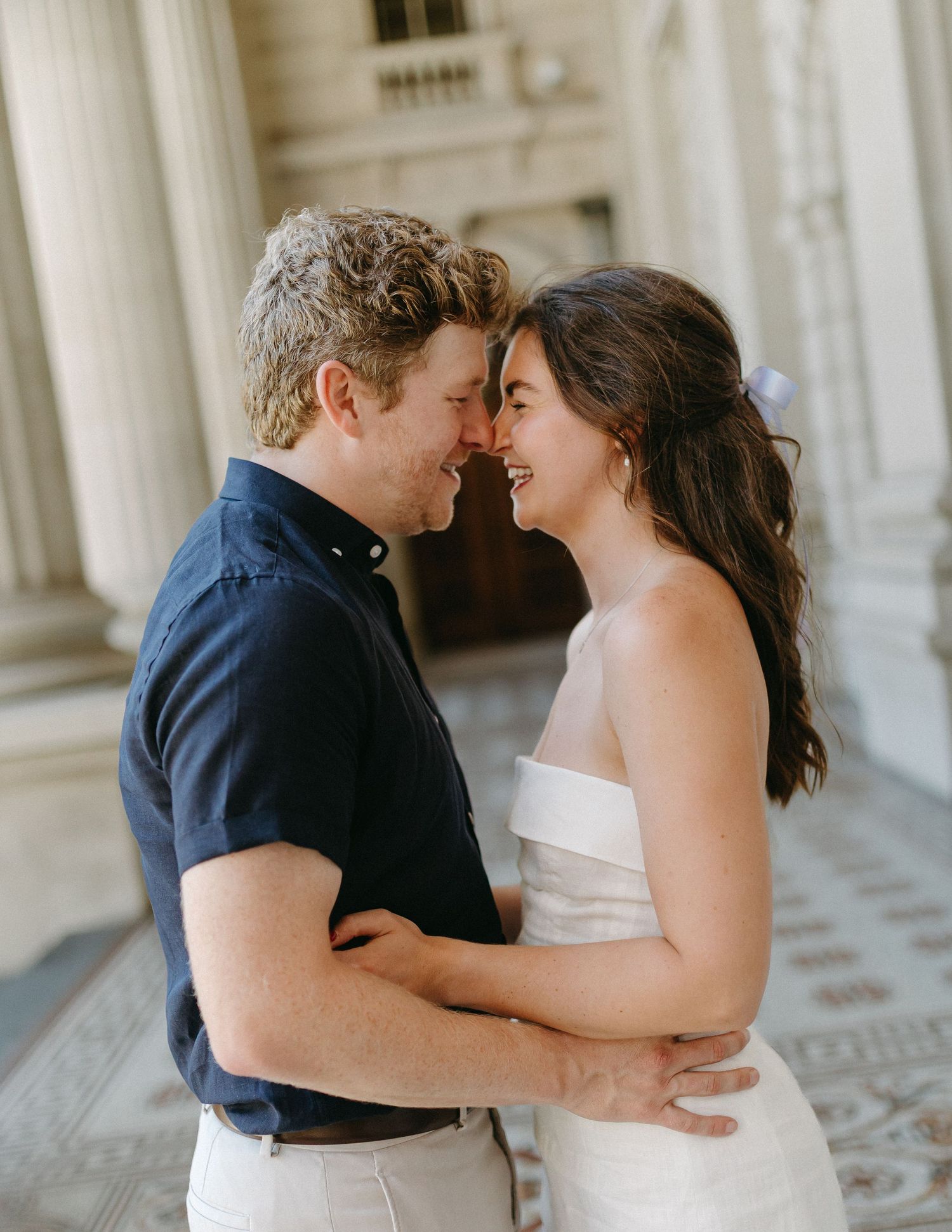 Smiling couple embracing and touching foreheads in front of classical white columns during engagement session.