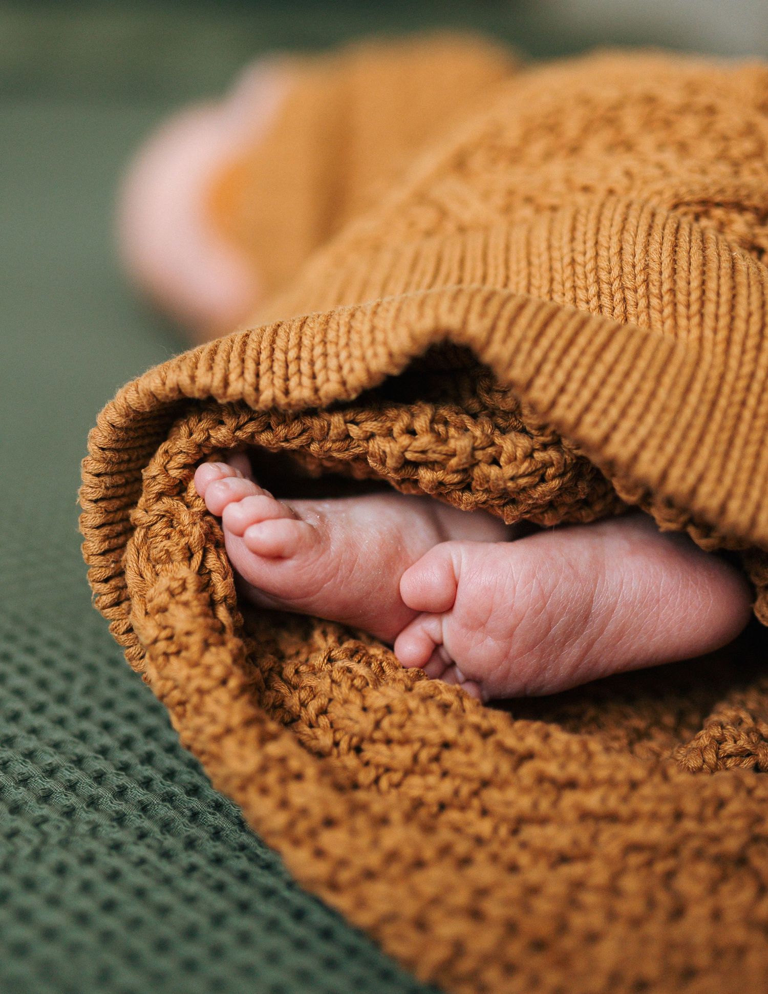 Close-up of tiny newborn baby feet peeking out from a mustard yellow knit blanket on a green surface.