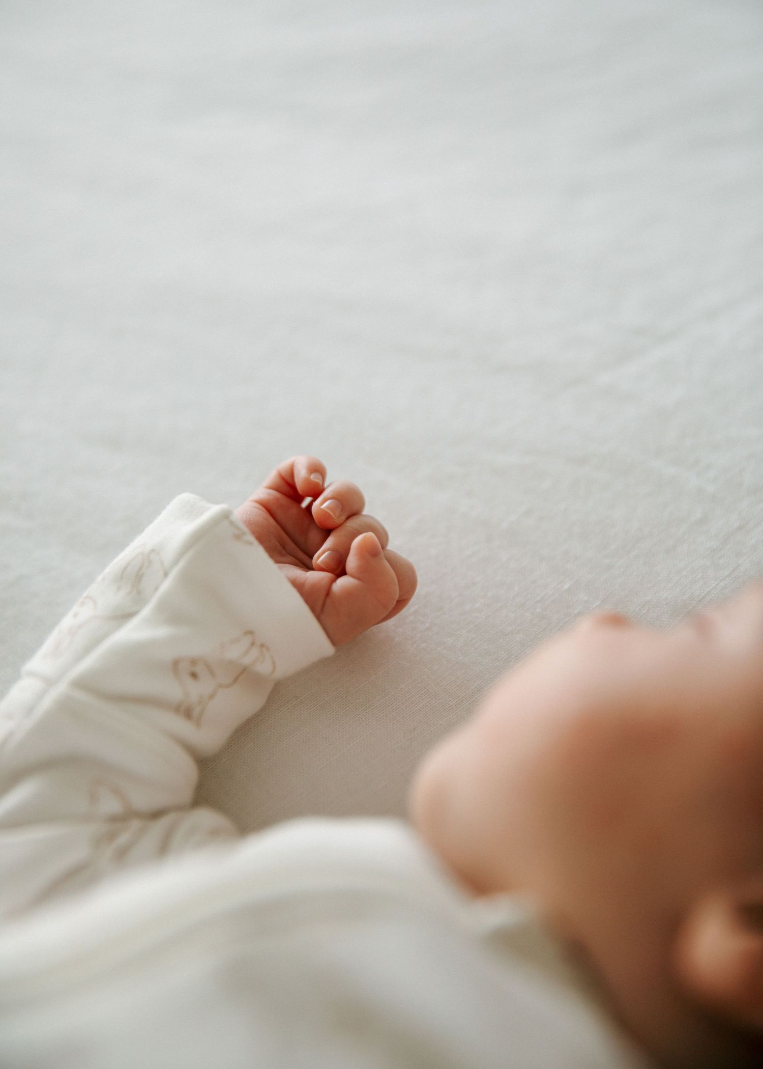 Newborn baby in white floral outfit lying on soft blanket with tiny hand raised.