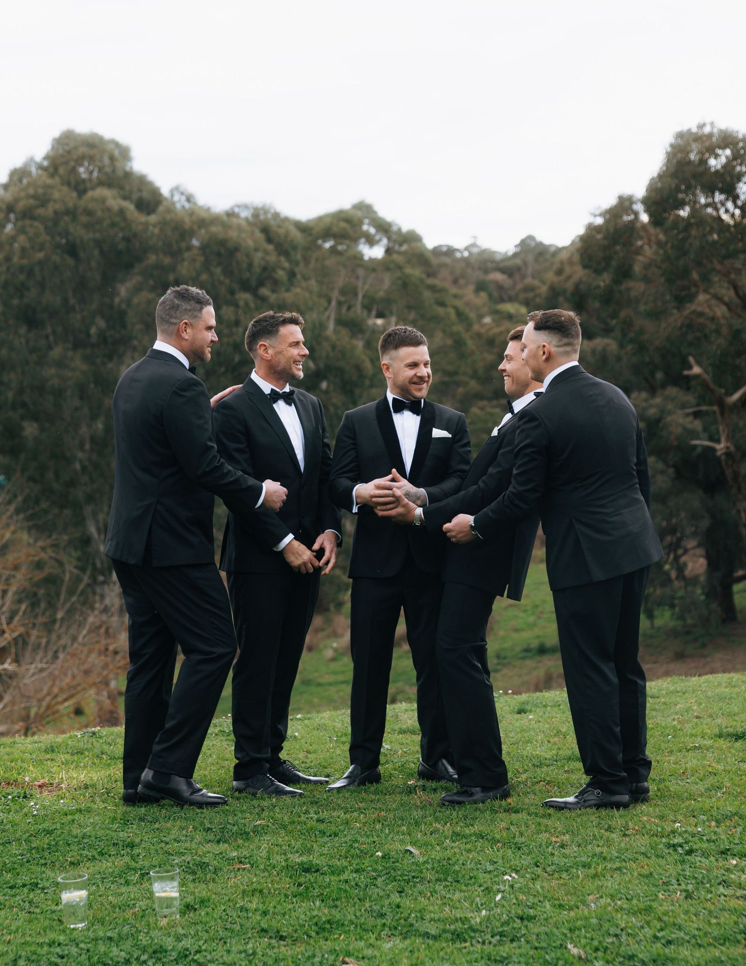 Four groomsmen in matching black tuxedos laughing together on green lawn outdoors.