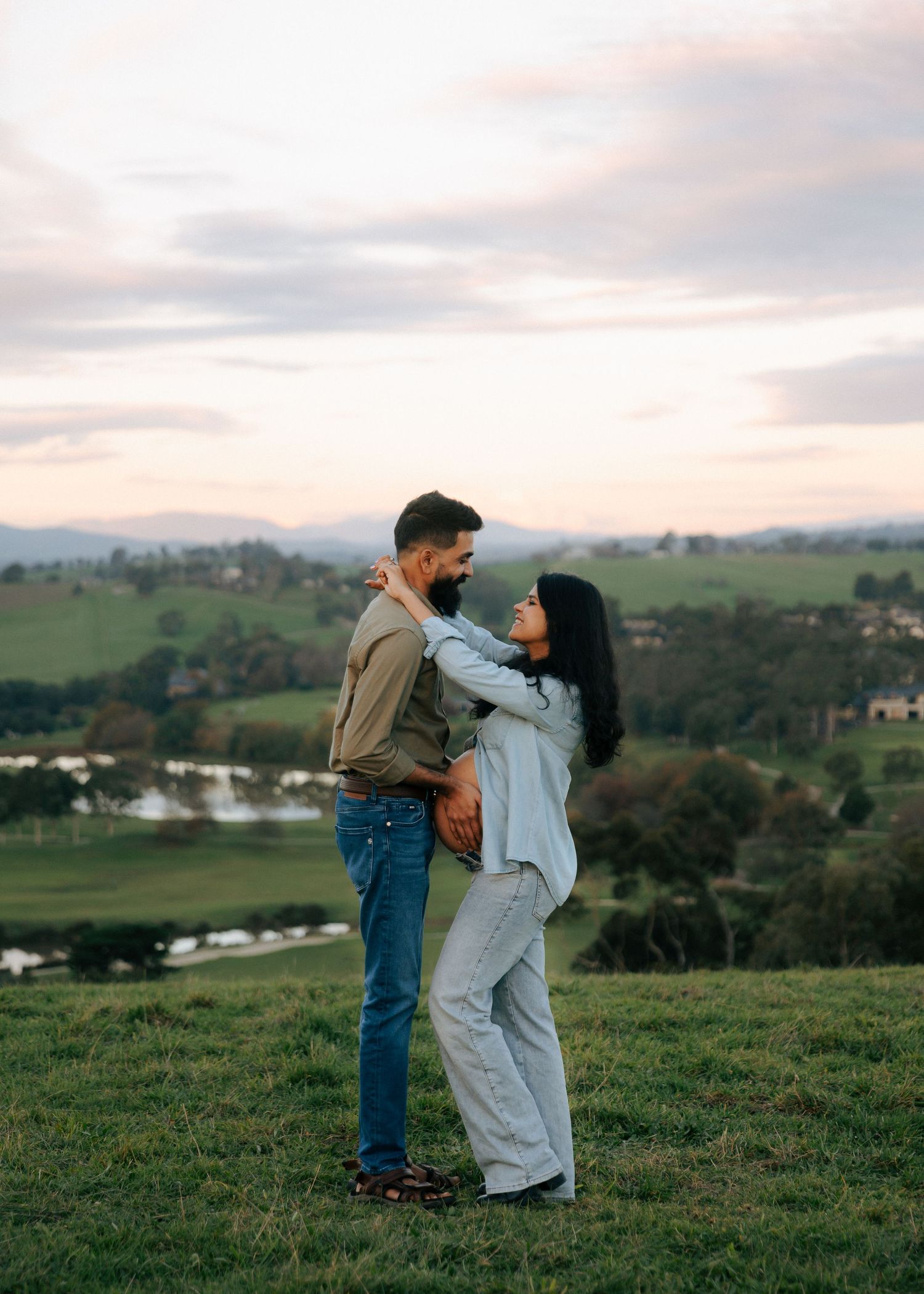 Couple embracing on a green hill at sunset with scenic countryside valley views behind them.