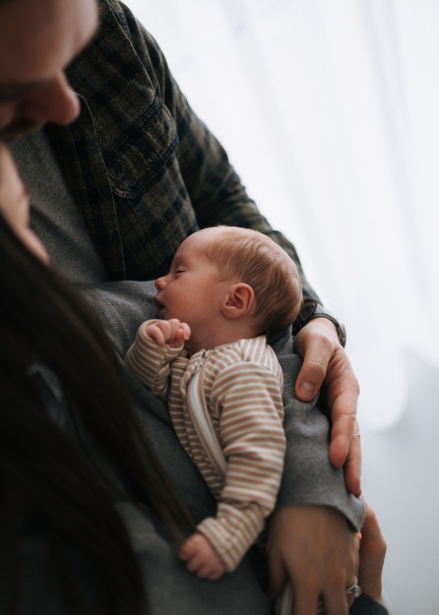 Close-up of a sleeping newborn baby dressed in a striped onesie being held by both parents.