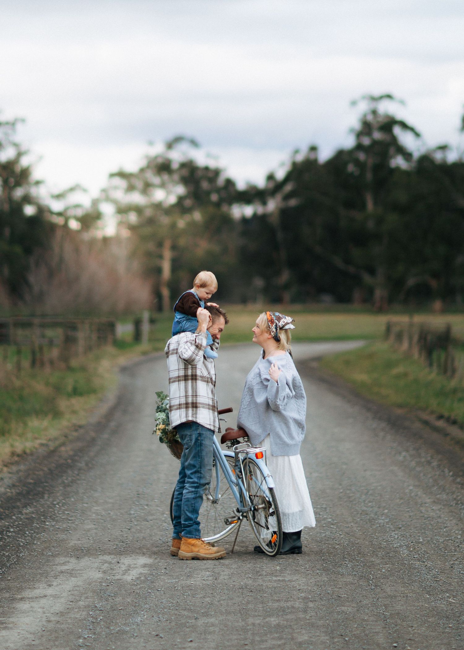 Family of three on a rural road, father holding toddler on shoulders, mother in floral headband beside a vintage bicycle.