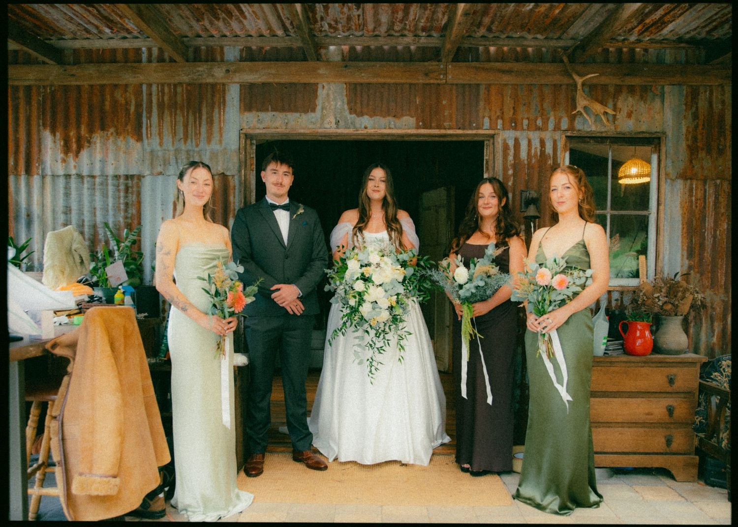 Bride and groom with bridesmaids in green dresses posing inside a rustic corrugated iron barn.