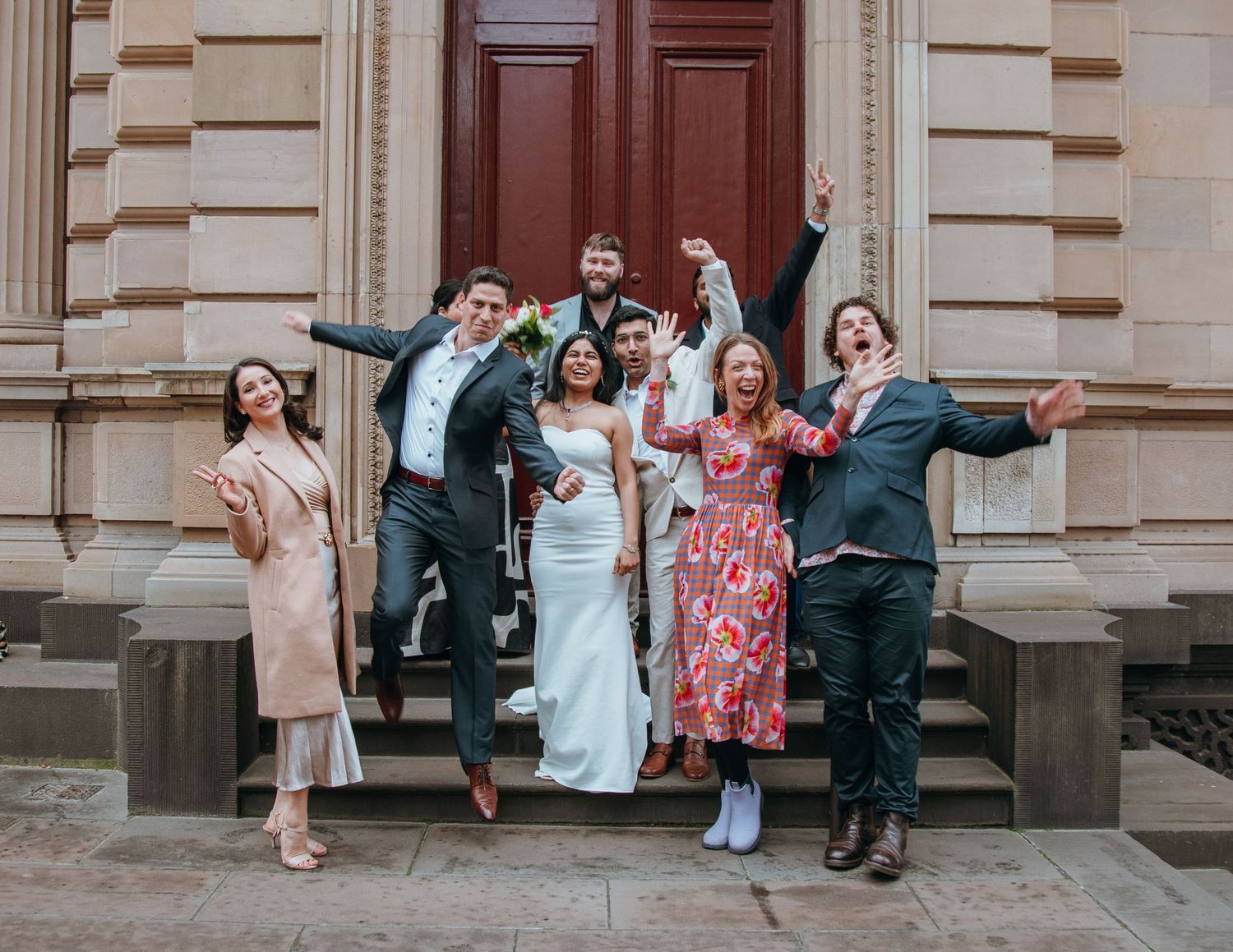 Joyful wedding party of six celebrates on stone steps outside grand building with arms raised in excitement.