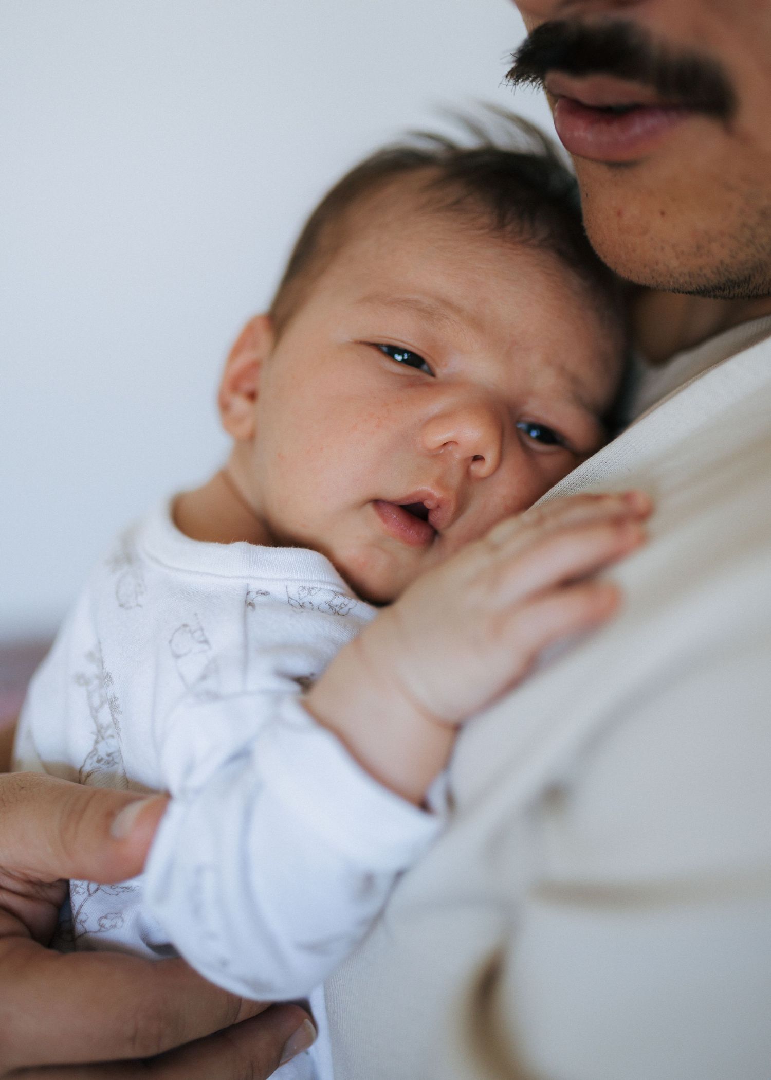 A newborn baby in a white printed onesie rests its head and tiny hand against the chest of a mustached man in a cream shirt.