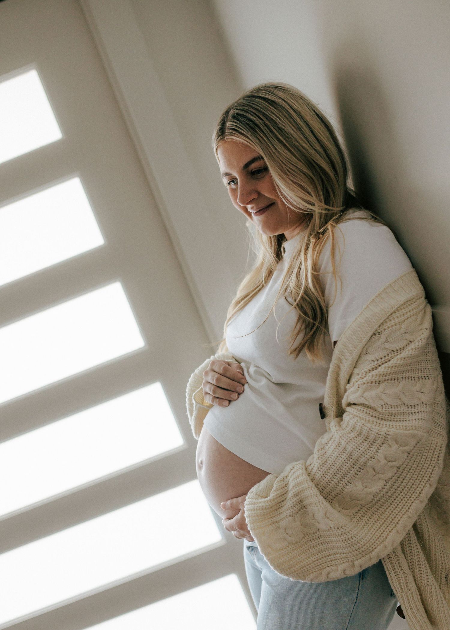 Smiling pregnant woman in white top and beige cardigan cradling baby bump by window with shutters.