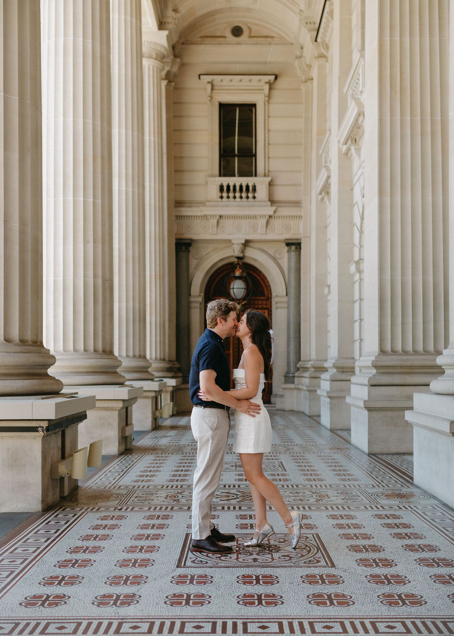 Couple kissing between grand white columns on ornate tiled floor during city engagement photoshoot.