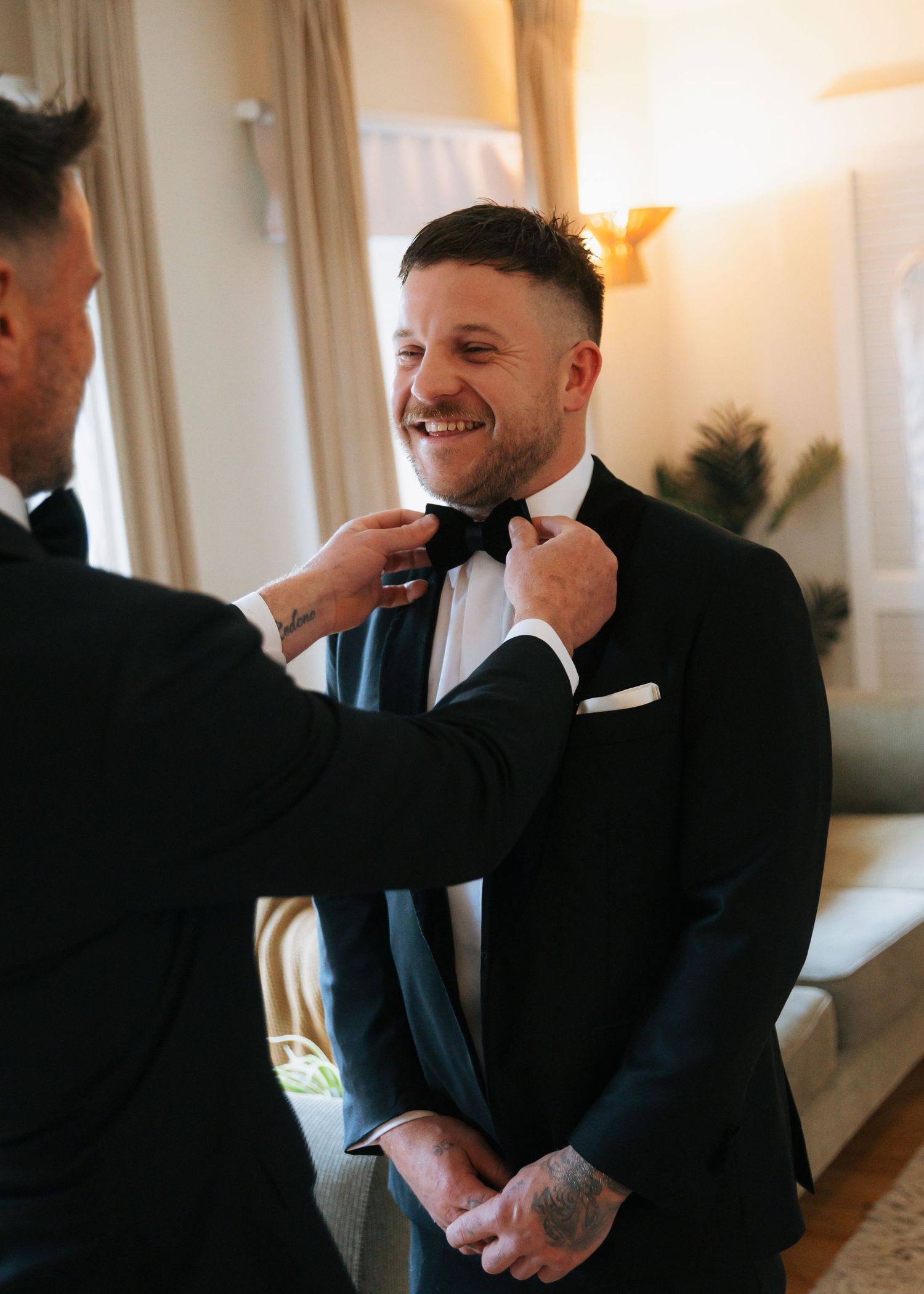 Groomsman adjusts bow tie of smiling groom in classic black tuxedo before wedding.