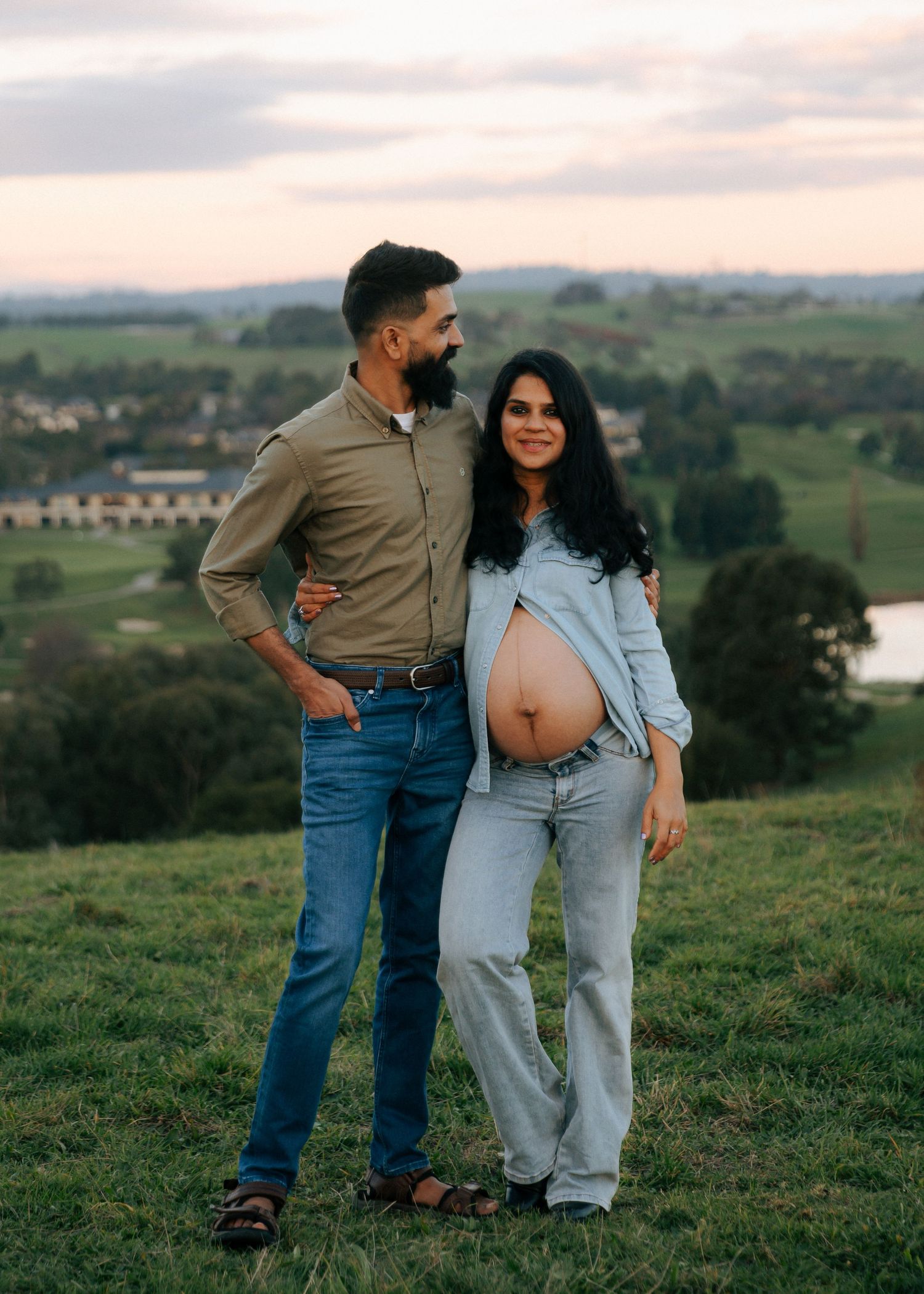 Couple poses together in green hillside landscape during outdoor maternity photoshoot.