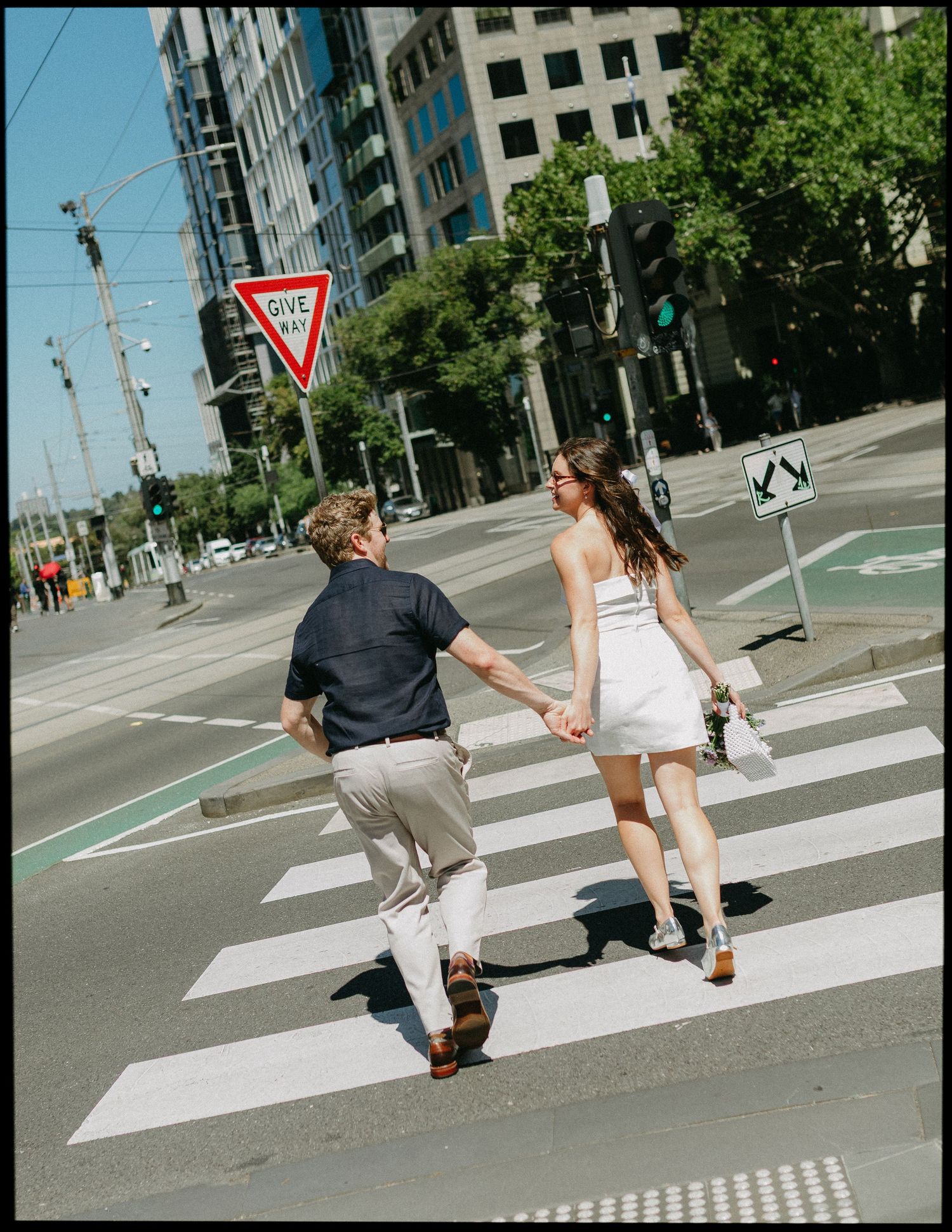 Newlywed couple crosses city street holding hands, bride in white mini dress with bouquet.