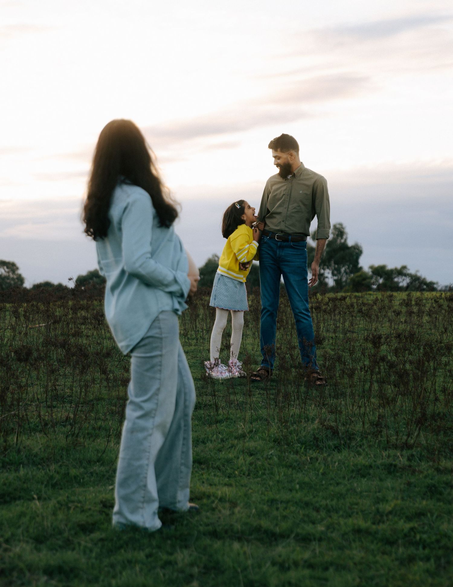 Family maternity photo with pregnant mother, father, and child in green field at dusk.