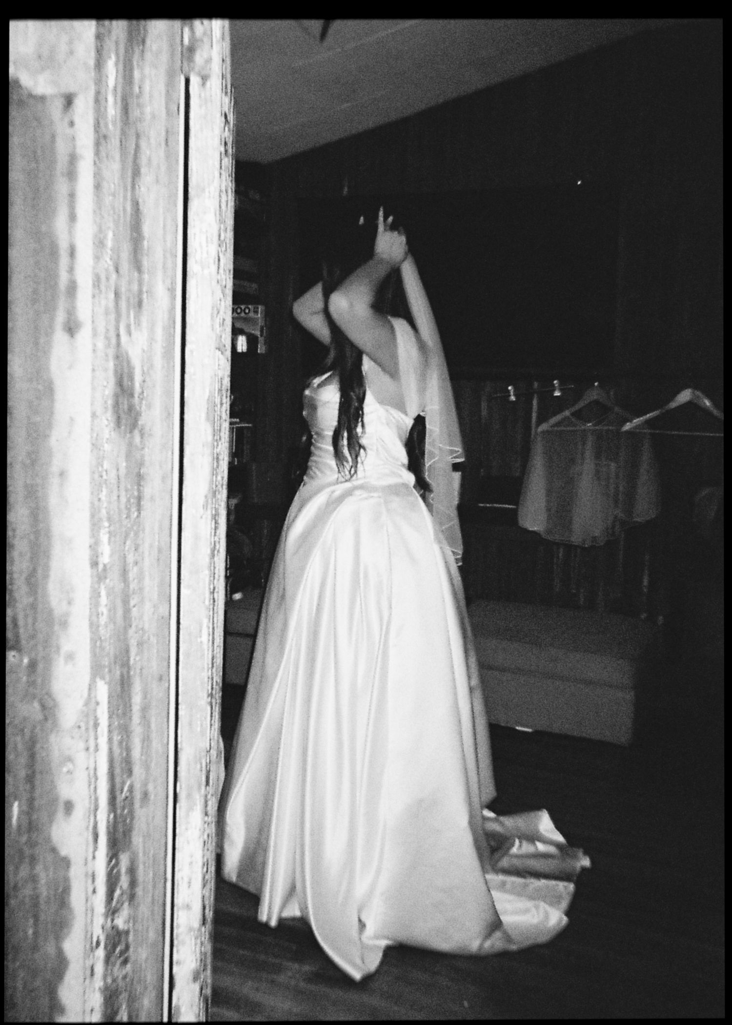Black and white photo of bride in strapless ballgown adjusting veil in a rustic barn dressing room.