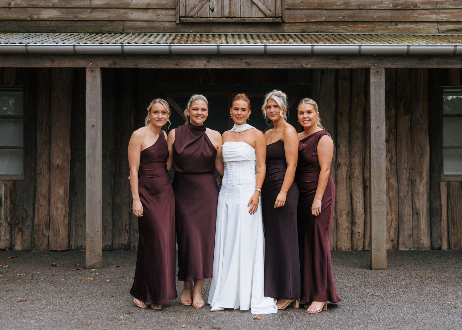 Bride with four bridesmaids in burgundy gowns outside rustic wooden venue.