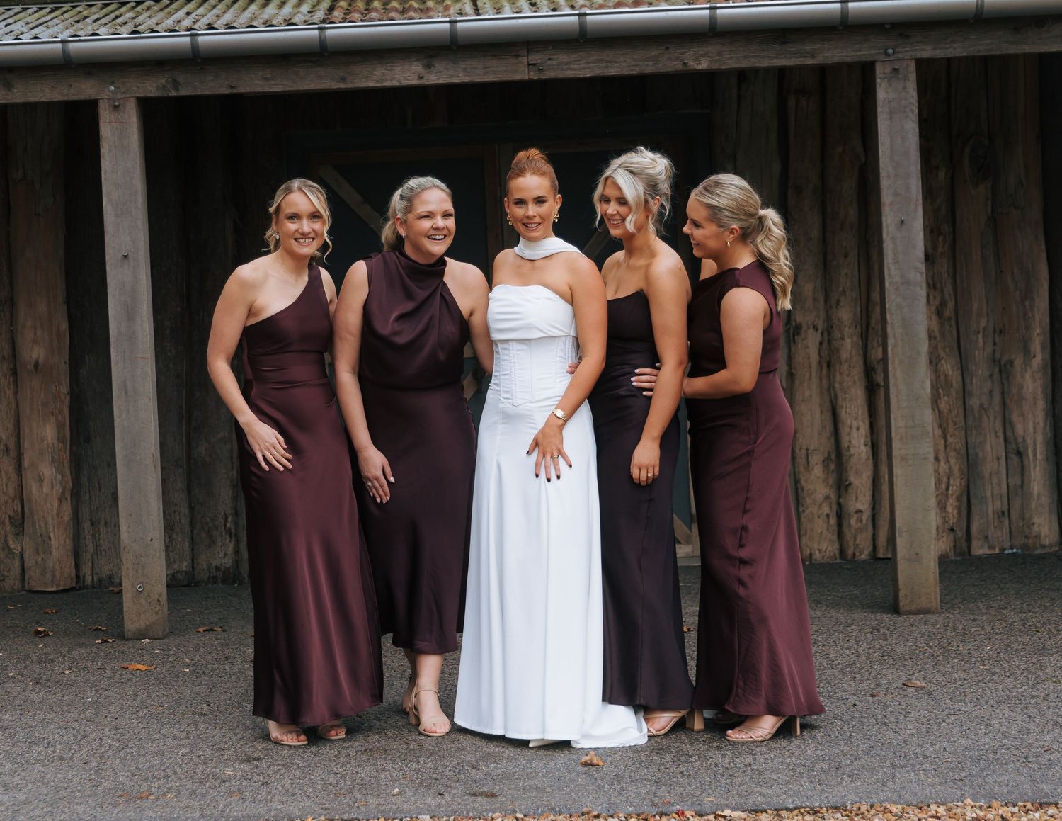 Bride in white gown smiles with four bridesmaids wearing dark burgundy dresses outside rustic wooden building.