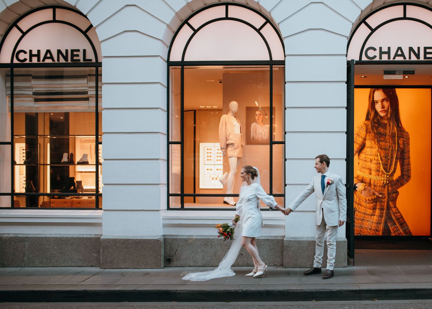 Bride and groom holding hands walking past illuminated Chanel boutique storefront at dusk.